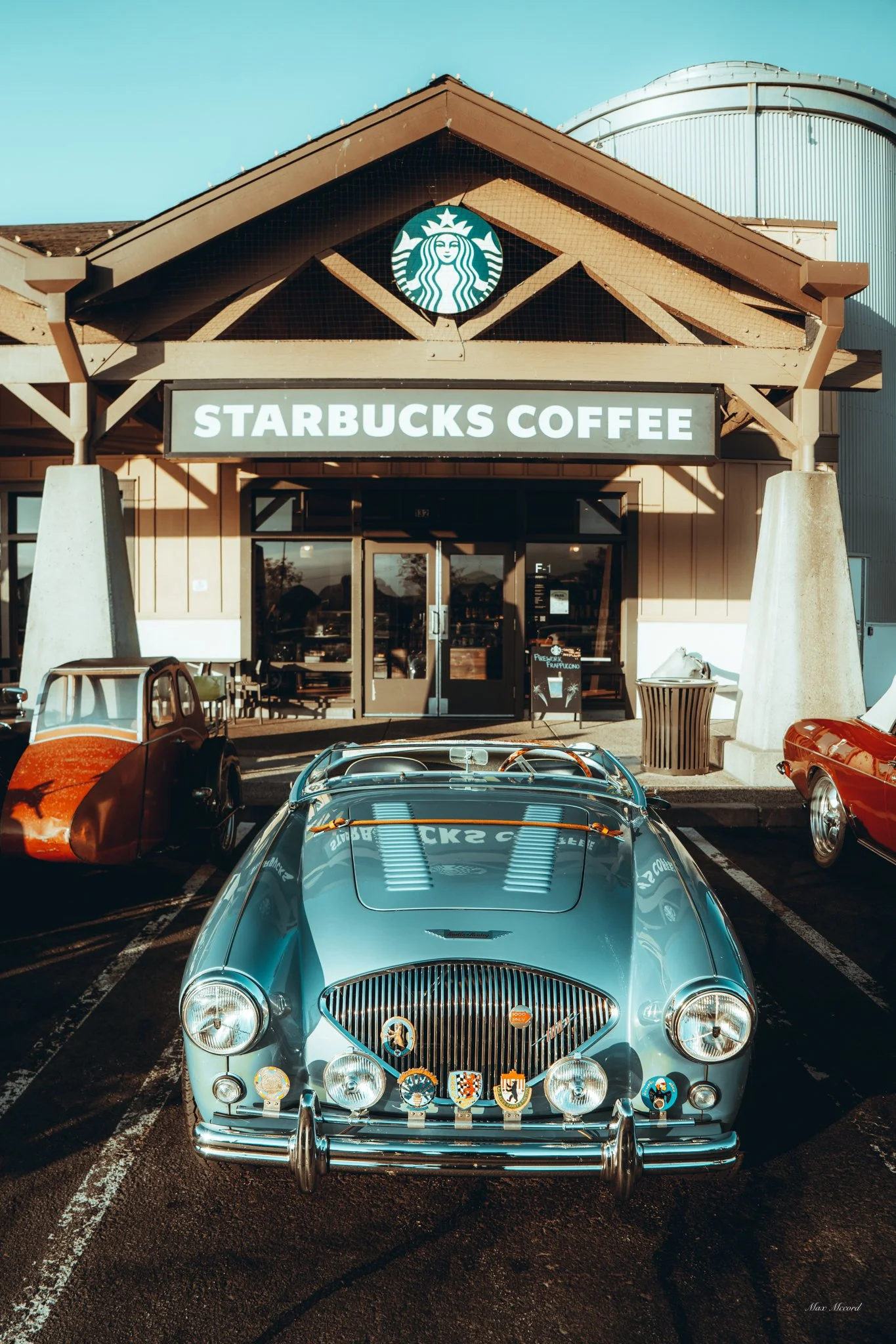 Vintage blue convertible car parked outside a Starbucks Coffee store, with classic cars on either side.