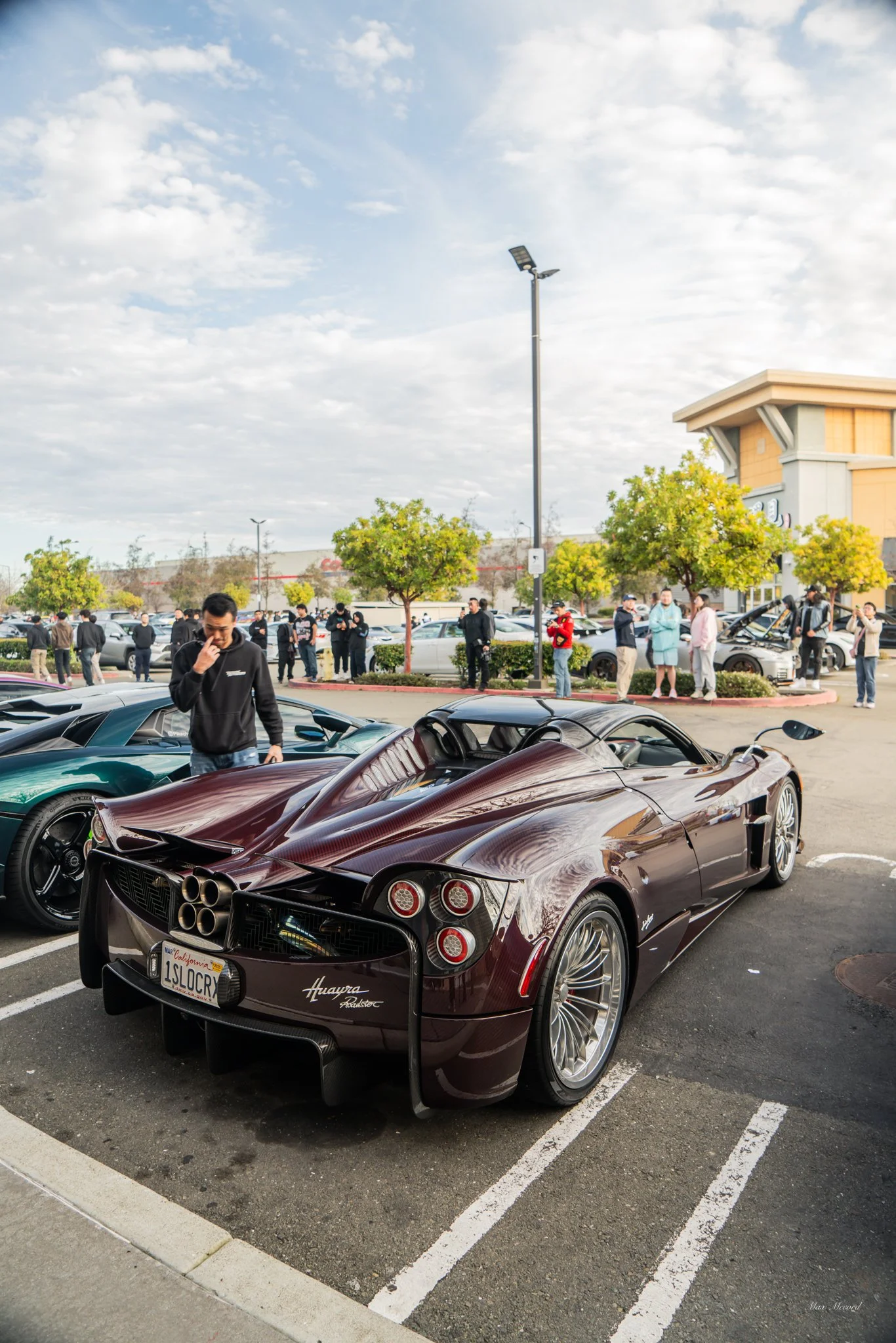 A dark red Pagani Huayra Roadster supercar parked in a parking lot during a car meet, with people looking at cars and trees in the background.