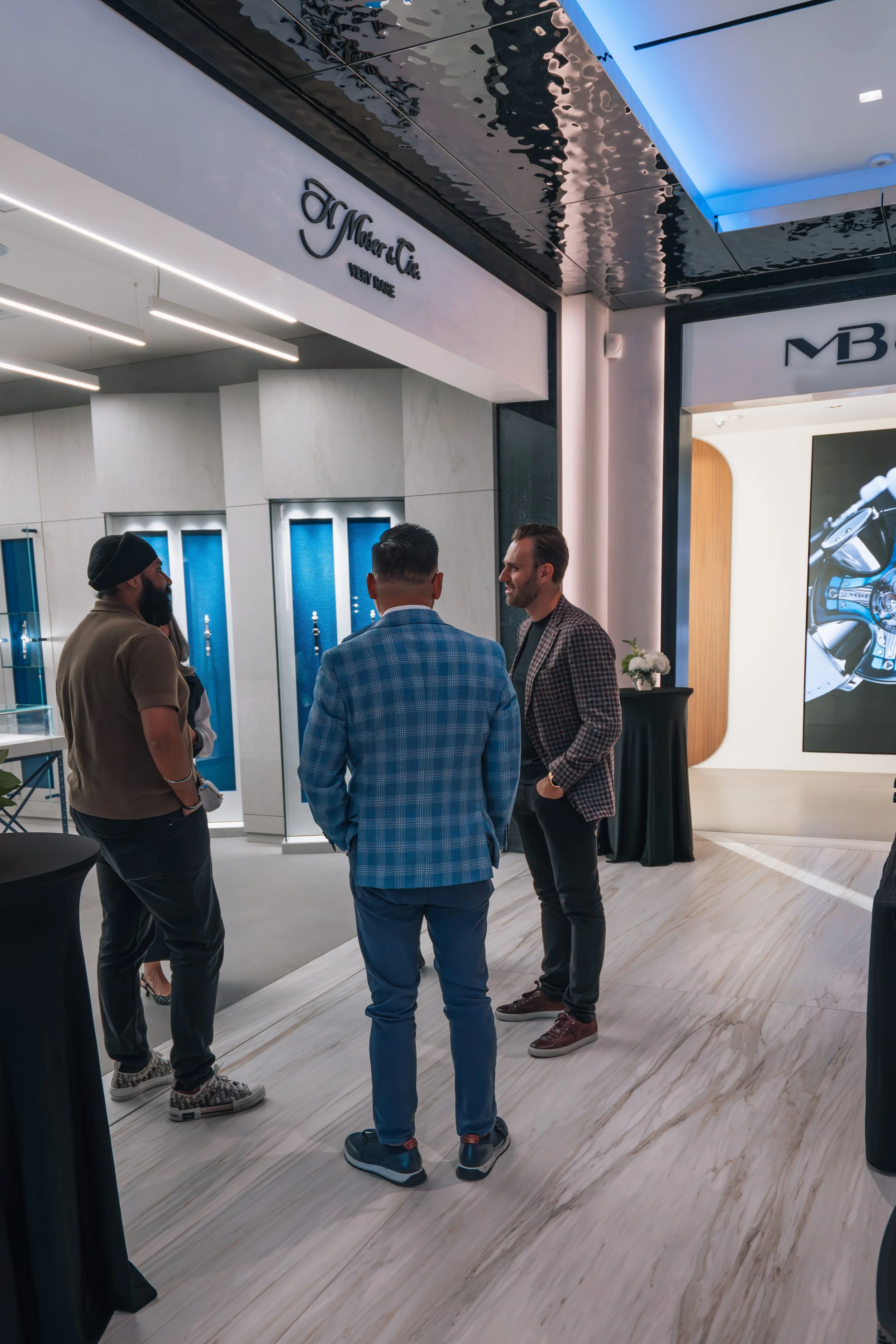 Three men are engaged in a conversation inside a modern showroom, with a large digital display and branding signage behind them.