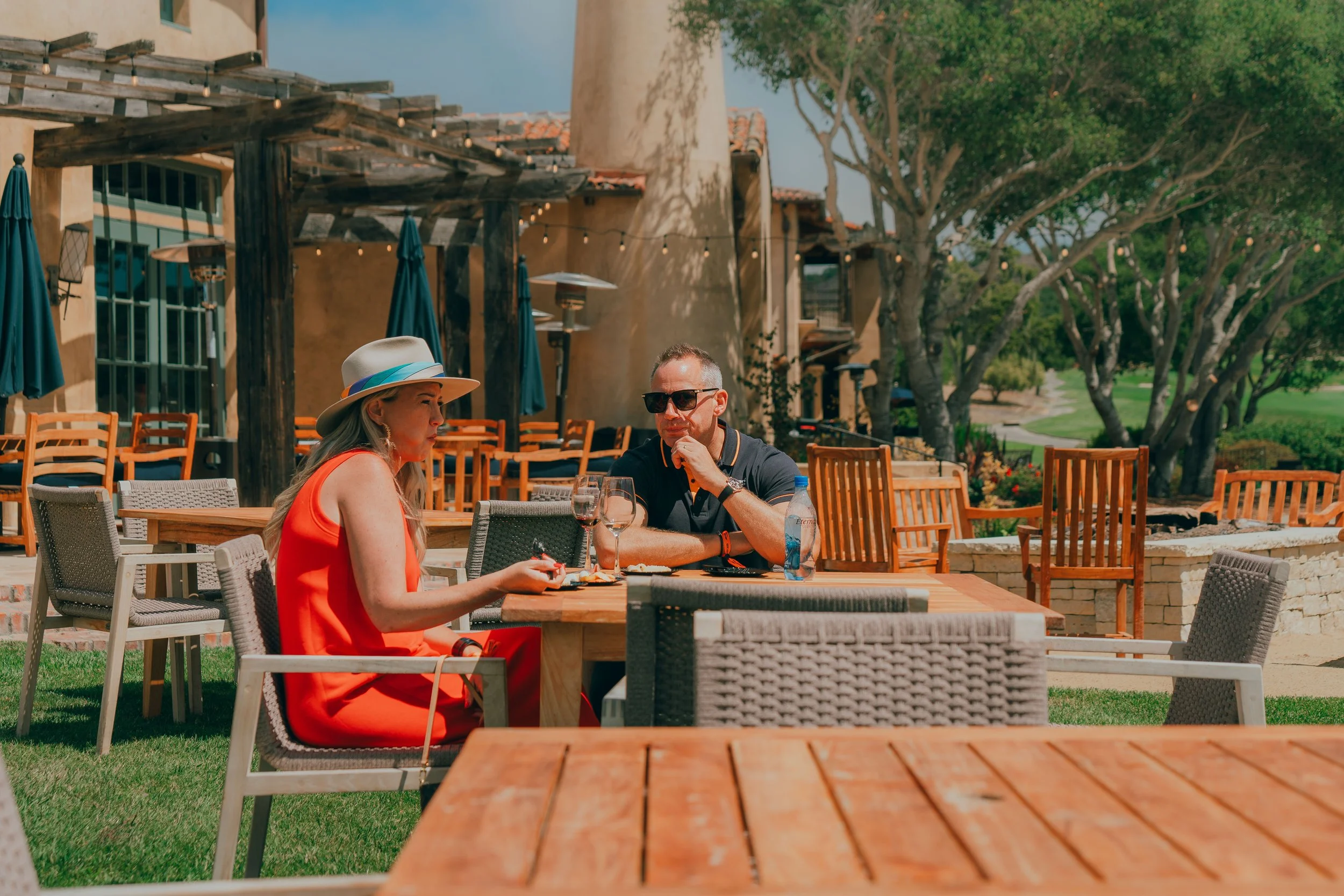 A woman and man sitting at an outdoor restaurant table, with empty chairs around, on a sunny day with trees and a building in the background.