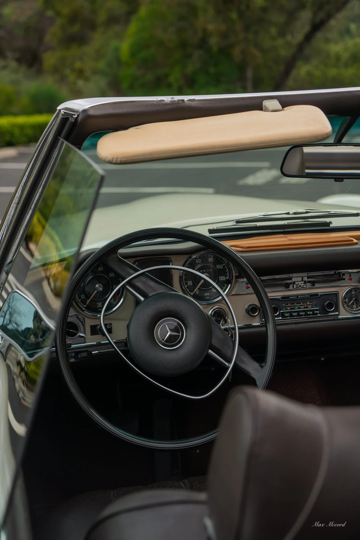 Interior of a vintage Mercedes-Benz convertible car, showing the black steering wheel with Mercedes emblem, dashboard with gauges and radio, and a view of the windshield and rearview mirror.