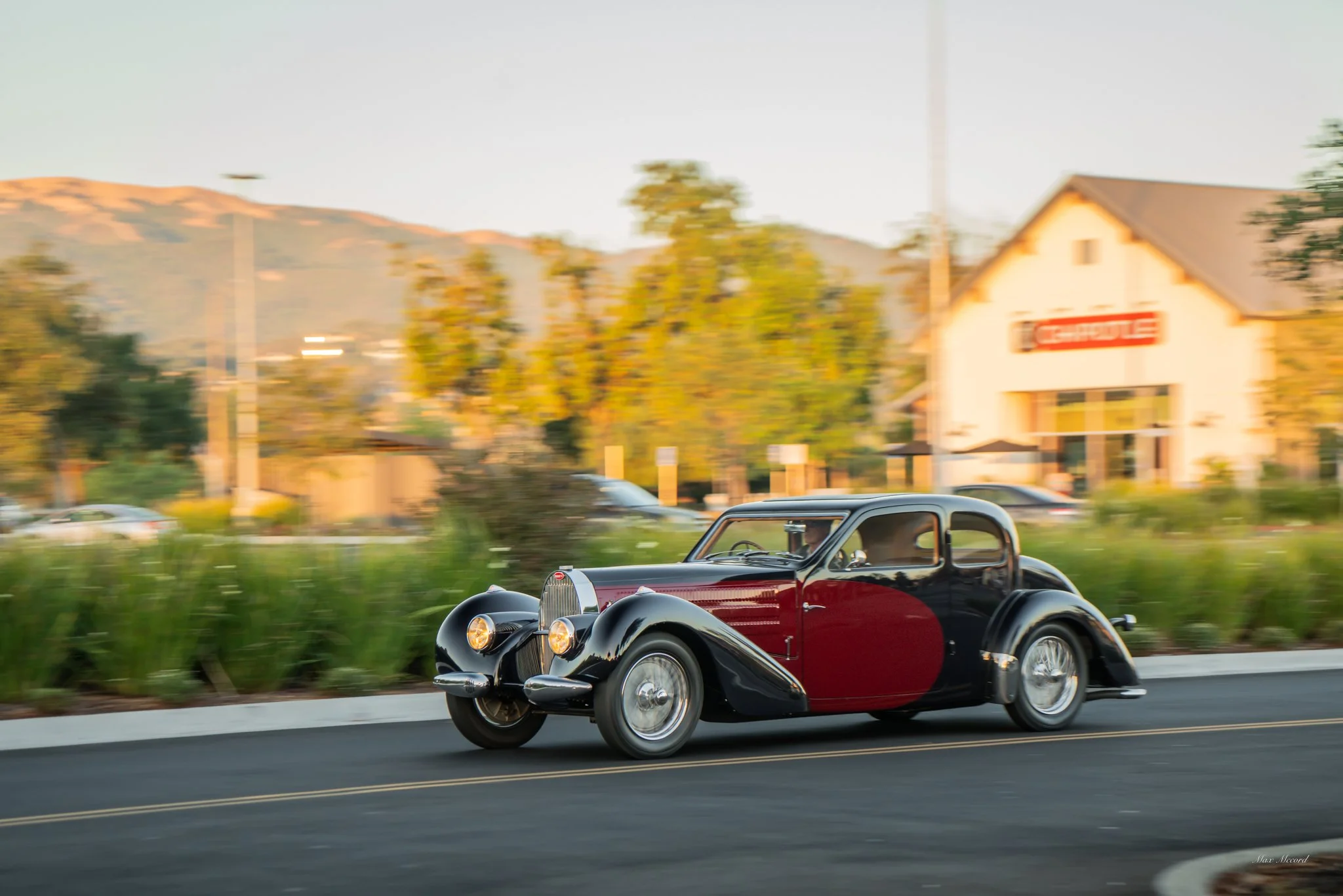 A vintage black and red classic car driving on a road with trees and a building in the background, with motion blur indicating speed.