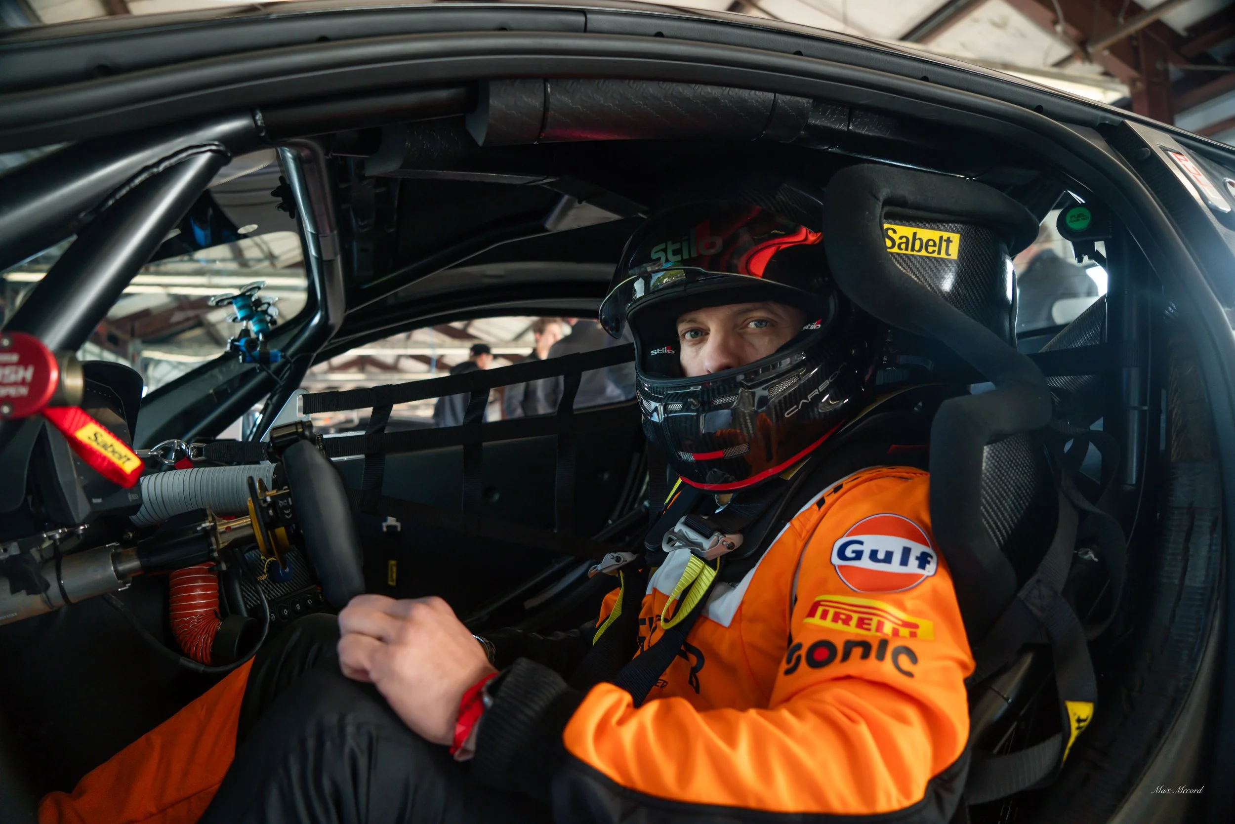 Race car driver sitting inside the cockpit of a race car, wearing an orange racing suit and a black helmet, with various racing sponsor logos visible on the suit and helmet.
