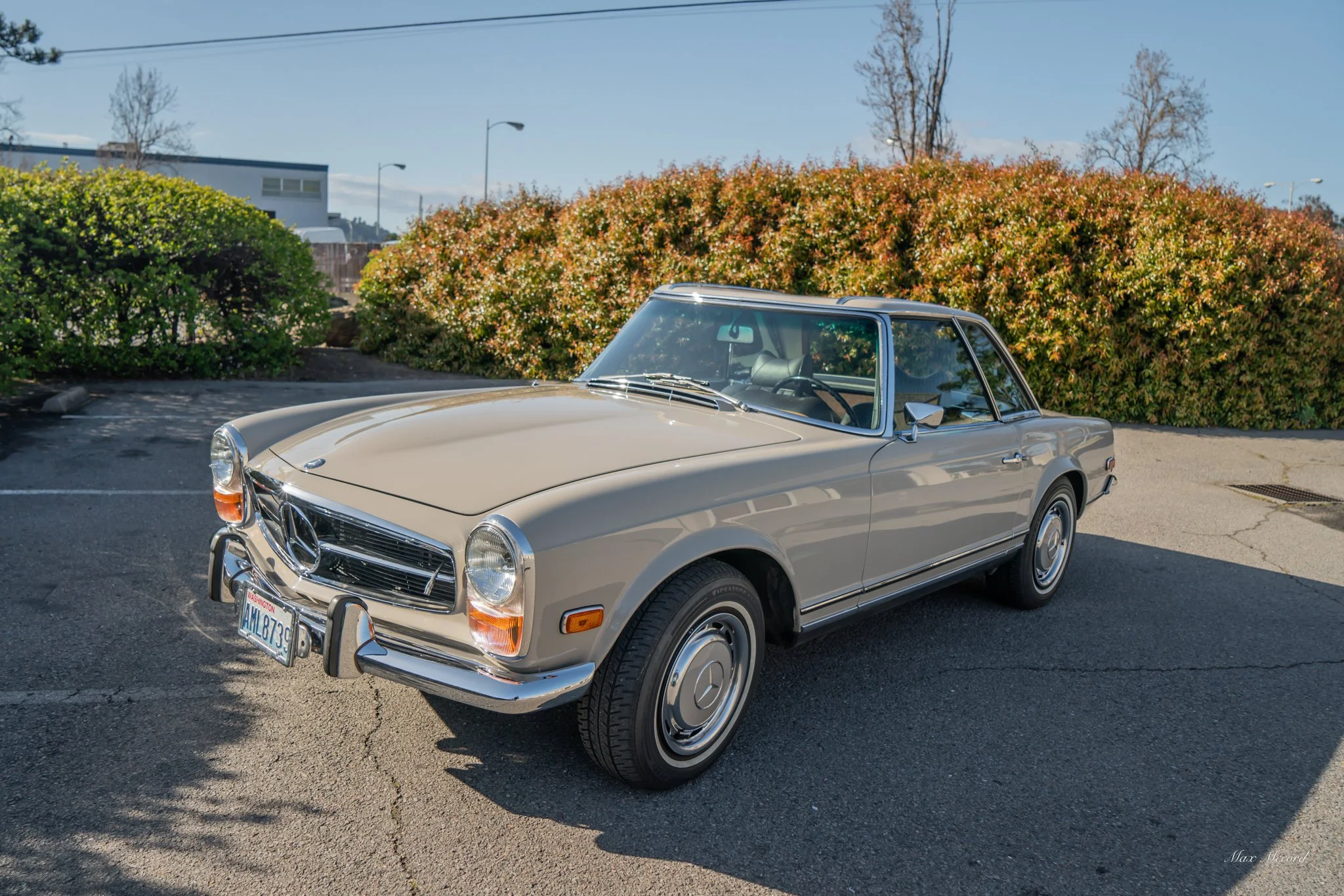 A vintage beige Mercedes-Benz car parked on a paved surface with green bushes and trees in the background under a clear blue sky.
