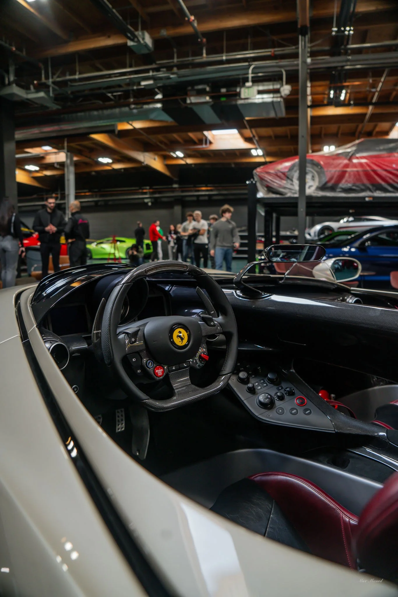 Inside view of a Ferrari's cockpit showing the steering wheel with Ferrari logo, dashboard, and control buttons, with a car exhibition or showroom and other luxury cars in the background.