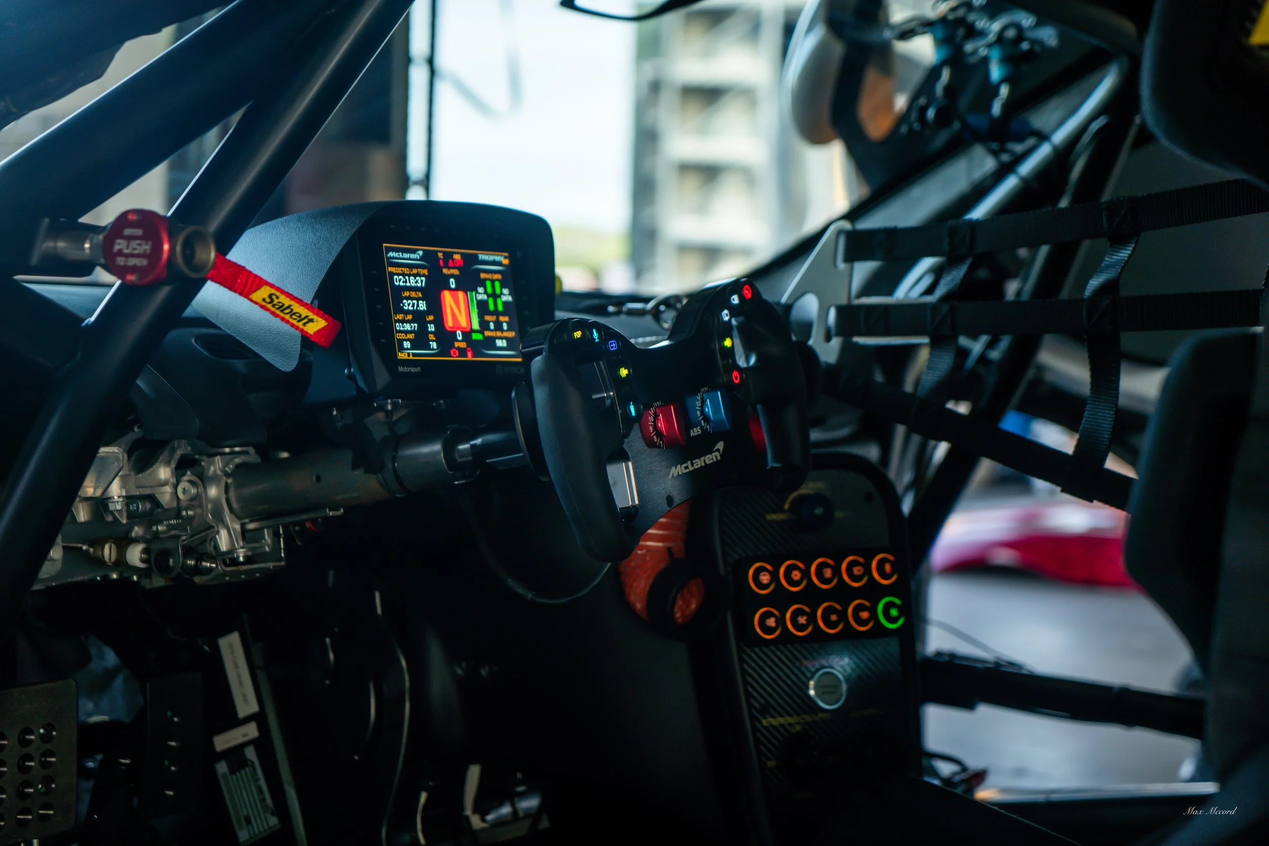 The interior of a racing car cockpit with a digital dashboard display, a racing steering wheel, and various control buttons and straps.