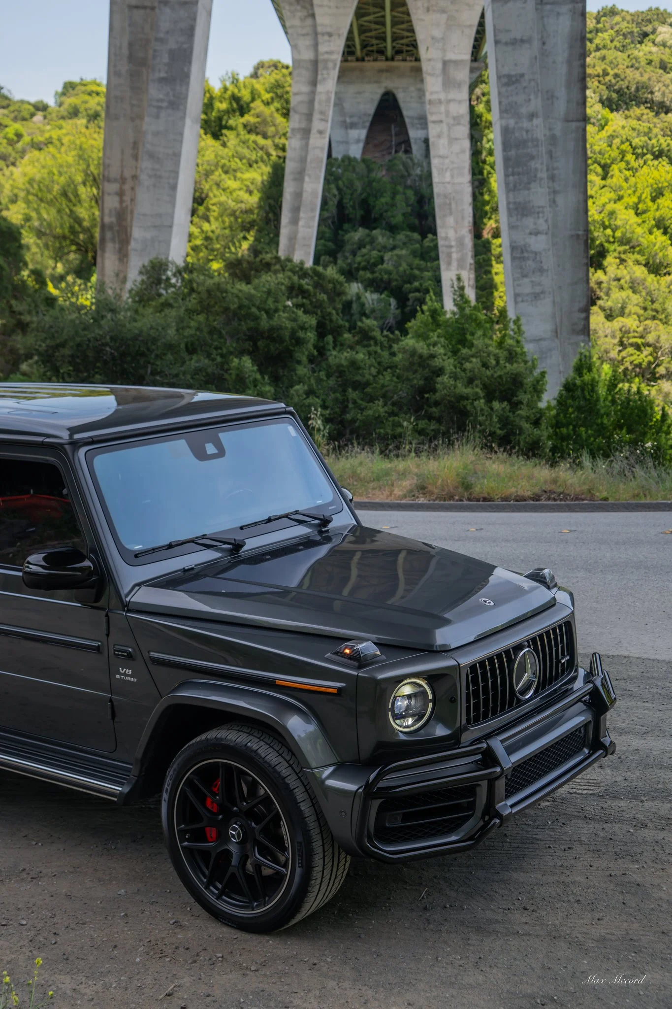A black Mercedes-Benz G-Class SUV parked on the side of a road with a large bridge structure and green forested hills in the background.