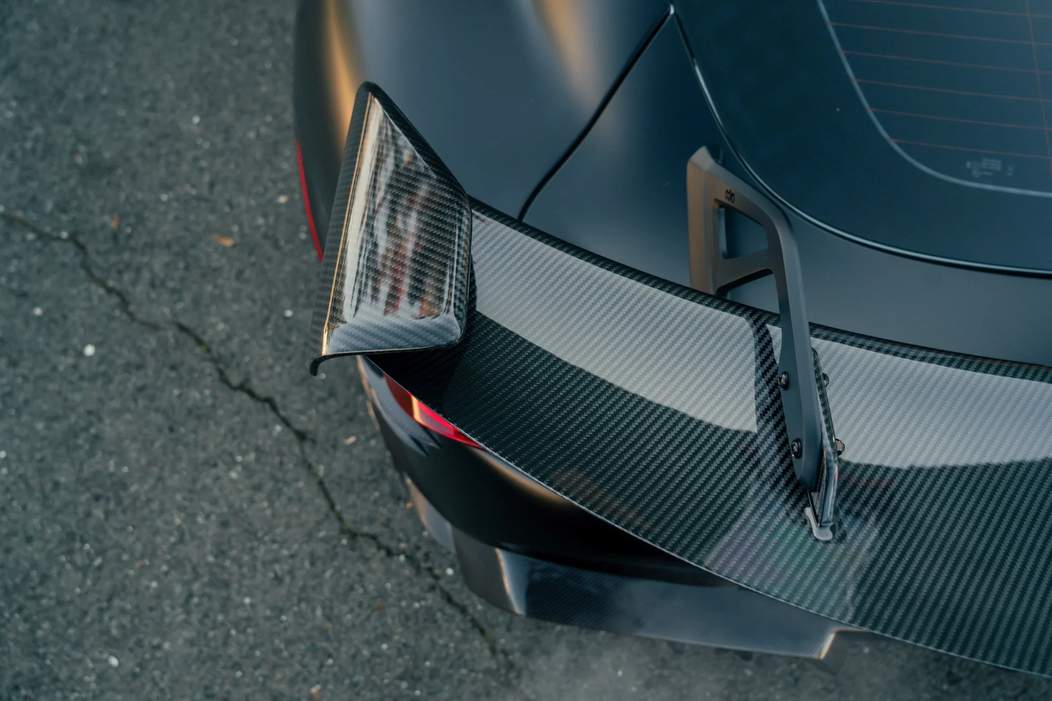 Close-up of the rear wing of a sports car, made of carbon fiber, mounted on the back of a matte black vehicle on a paved surface.