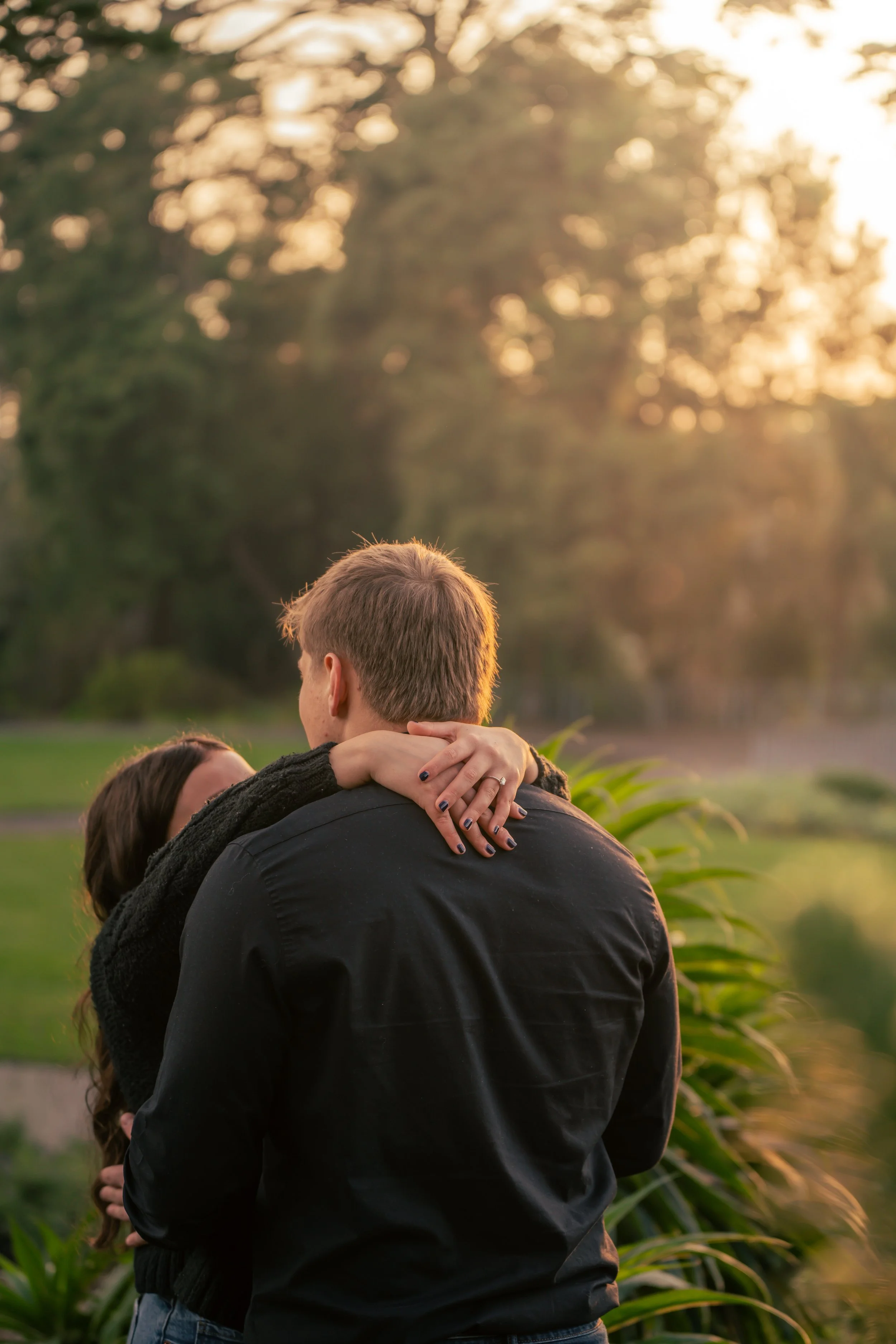 A couple embracing outdoors during sunset, with the woman’s arms around the man's neck and her face hidden. The scene is set in a green park with tall trees in the background.