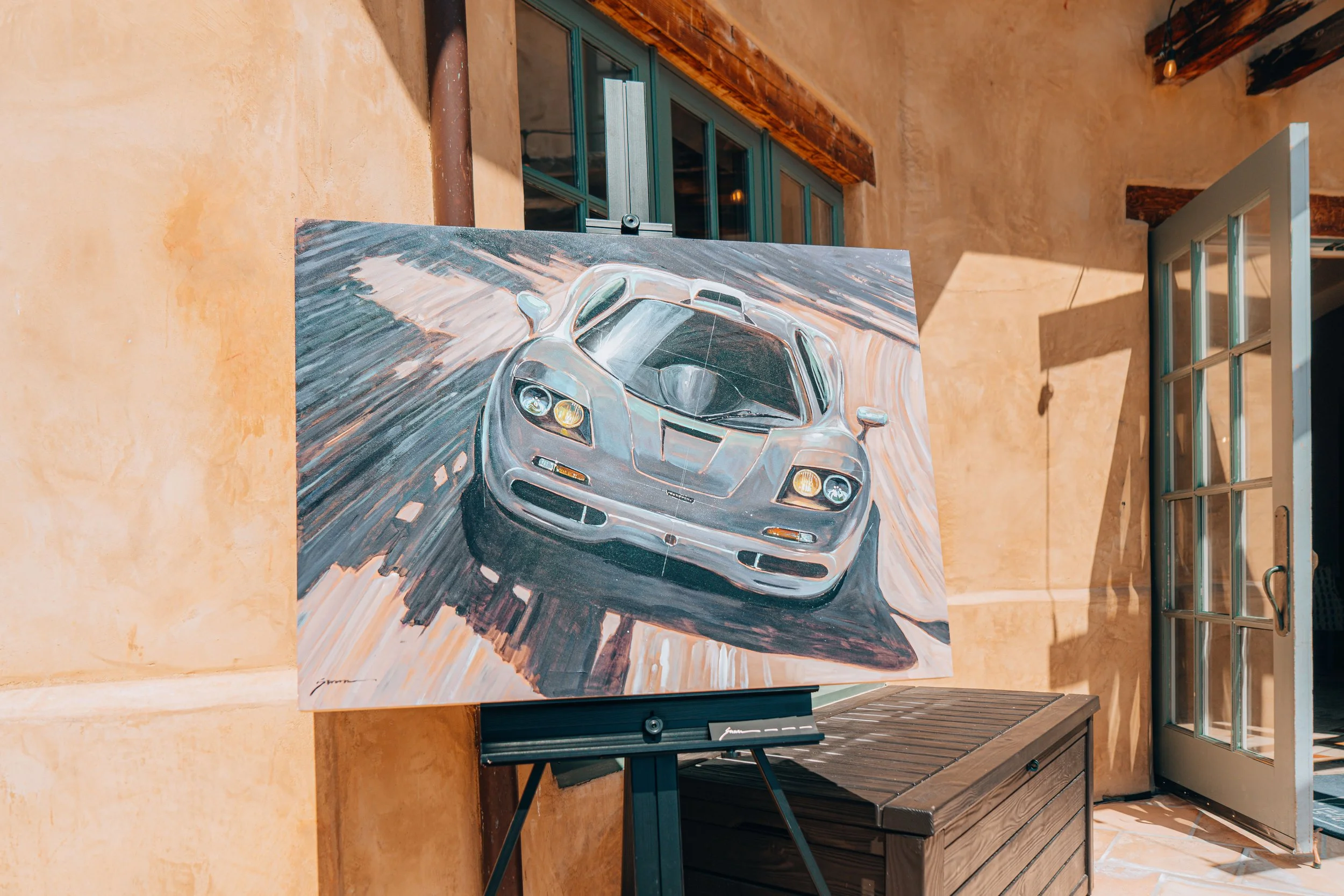 Painting of a silver sports car on an easel inside a room with beige walls and a glass door to the right.