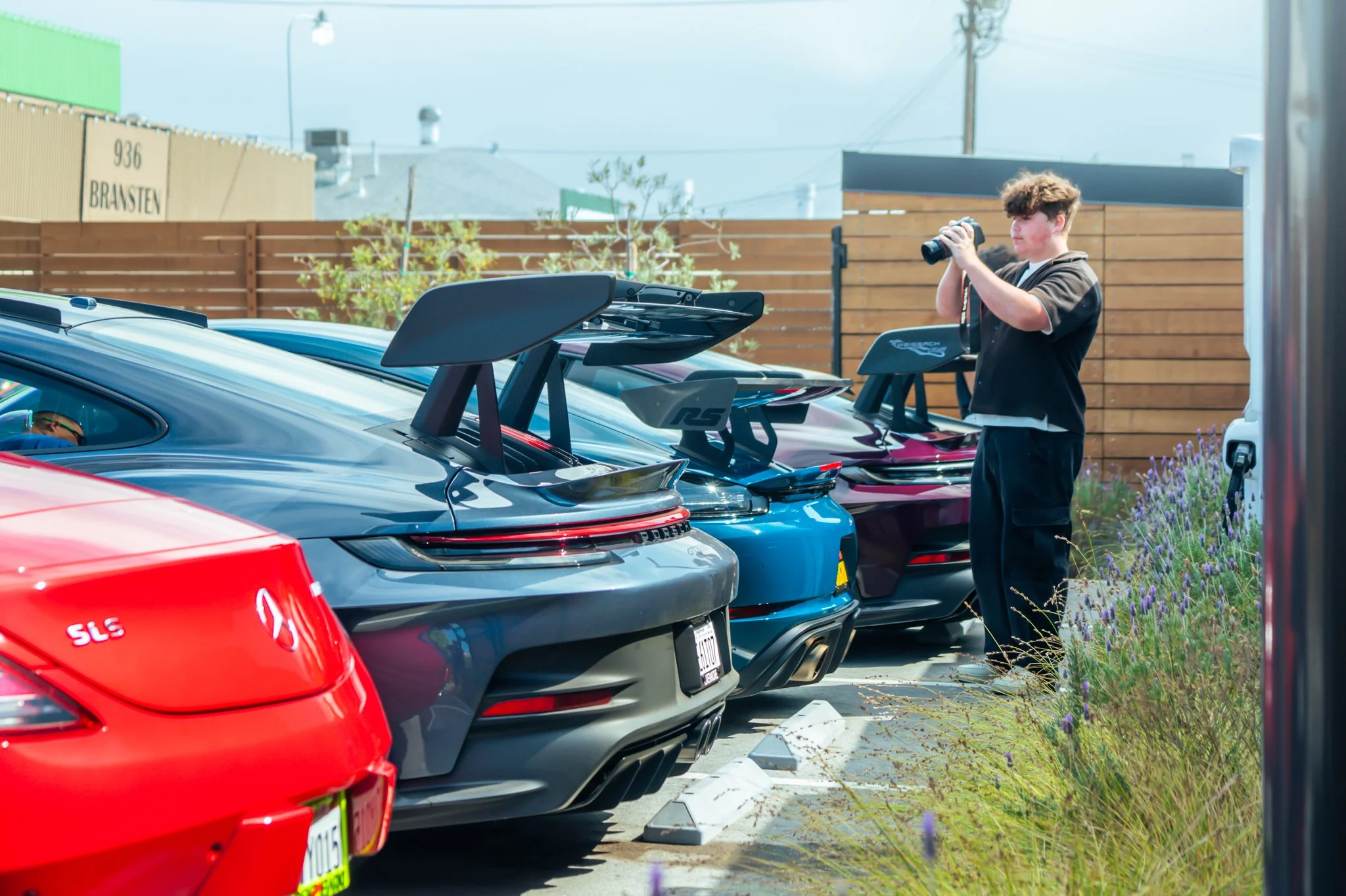 A young man with curly hair wearing a black t-shirt is standing next to a row of three sports cars with large rear spoilers, taking a photo with his smartphone. The cars are parked in a lot with a wooden fence and some greenery and purple flowers nea