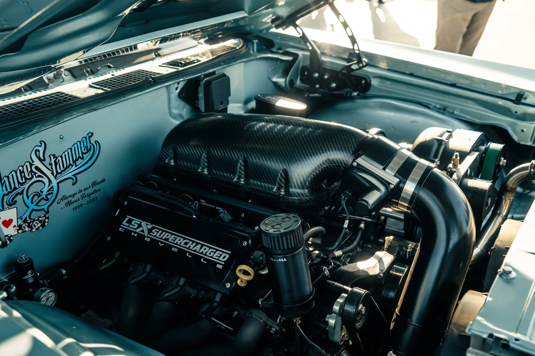 Close-up of a race car engine with black components, a carbon fiber air intake, LSX Supercharged Chevrolet engine cover, and various hoses and parts inside the car's engine bay, with a sticker that reads "Always on our Heart" and "1999-2022".