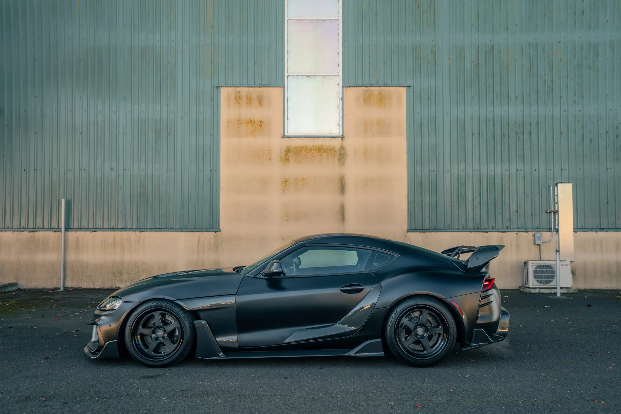 A sleek black sports car parked on a paved surface in front of a weathered green industrial building with a small window and air conditioning unit.