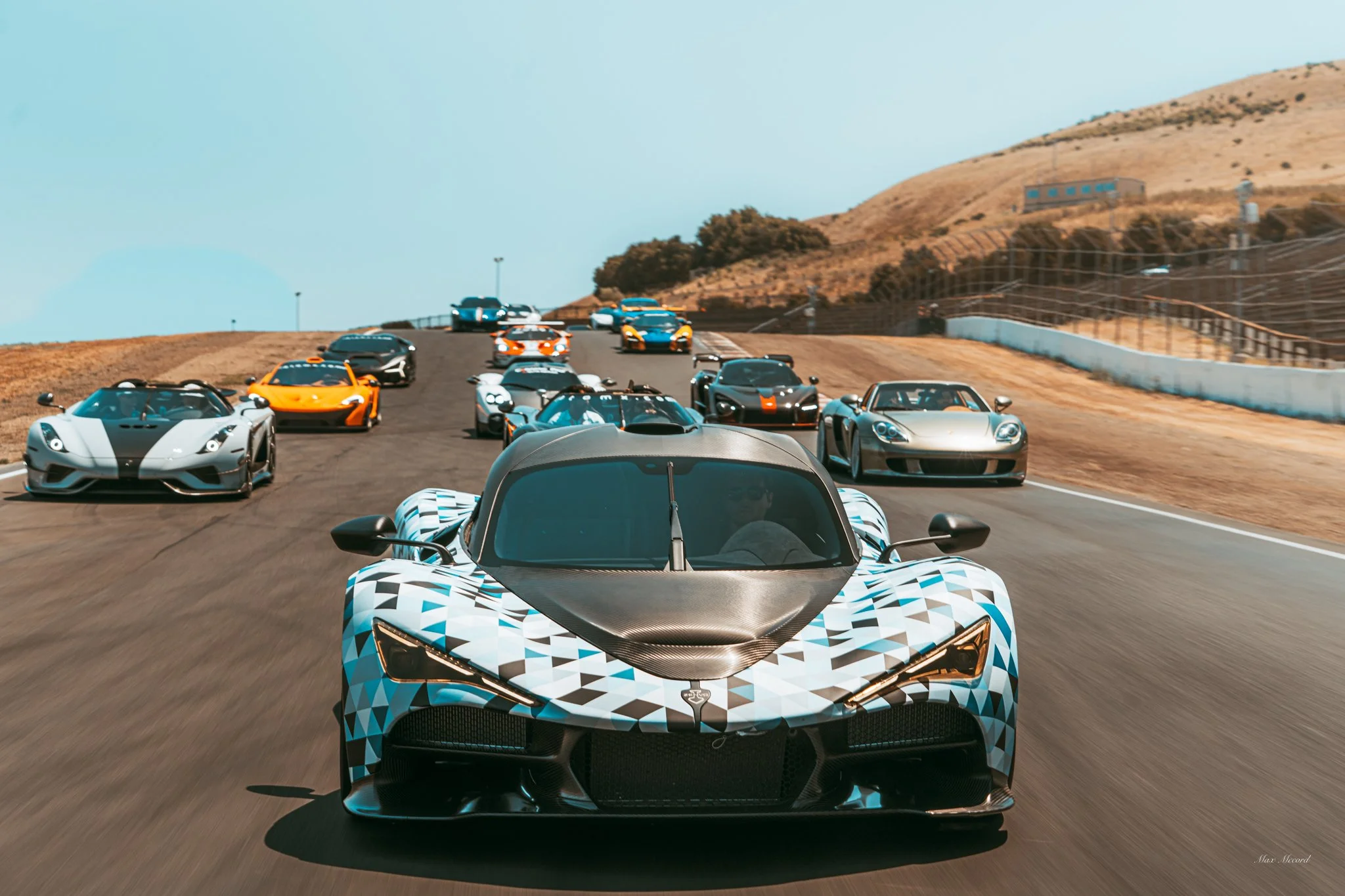 A lineup of high-performance sports cars on a racetrack, with a distinctive racing car in front, featuring a black and white geometric camouflage pattern, surrounded by various colorful supercars, and a hilly landscape in the background.