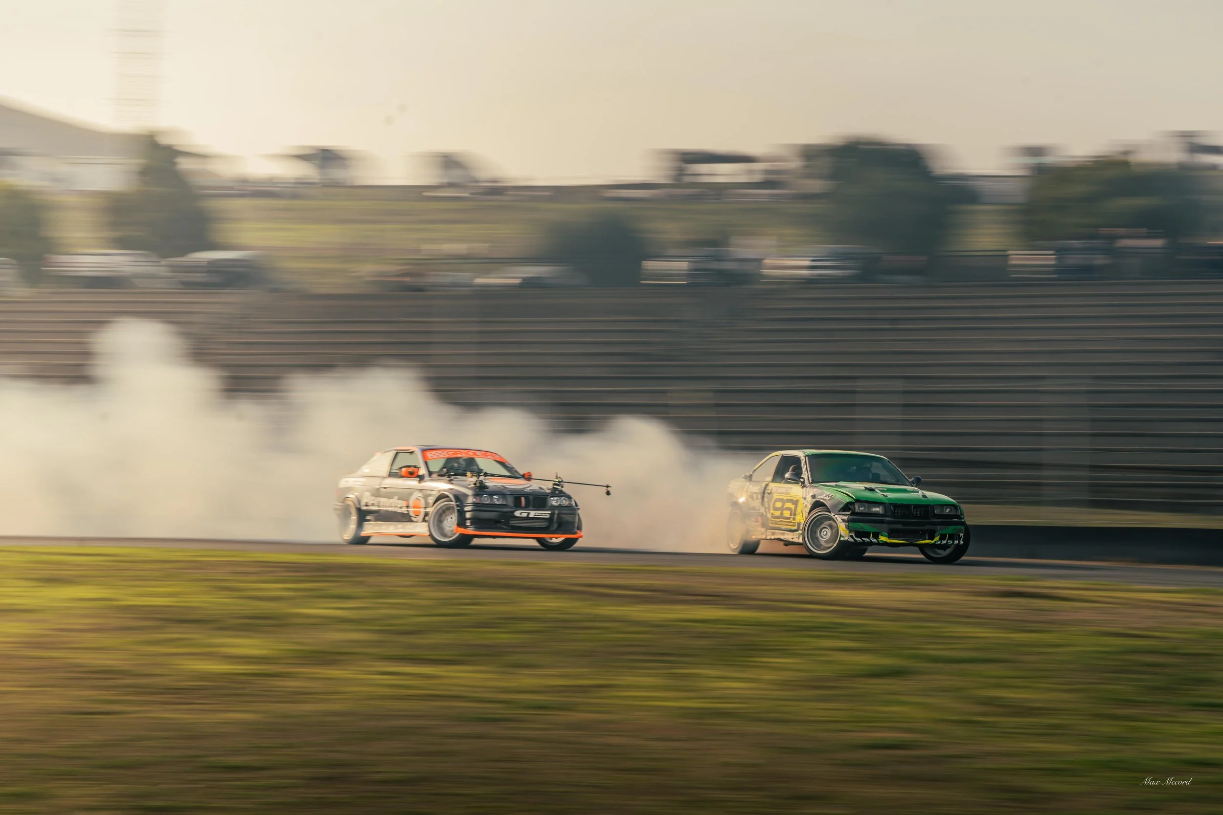 Two cars drifting on a race track with smoke clouds behind them, blurred background indicating high speed.