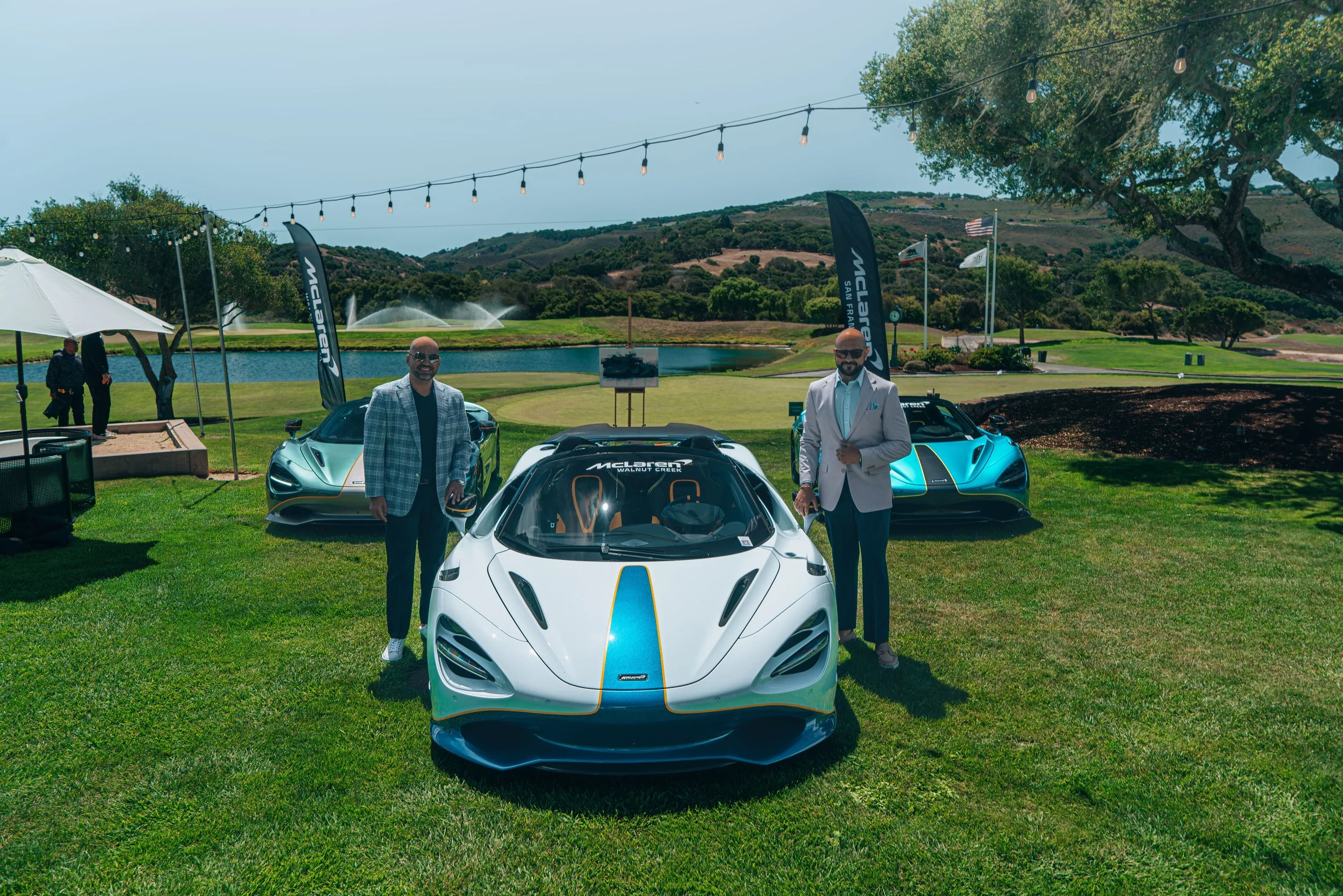 Two men in suits standing next to luxury sports cars on a grassy area near a lake, with a golf course and hills in the background, during a sunny day with string lights overhead.