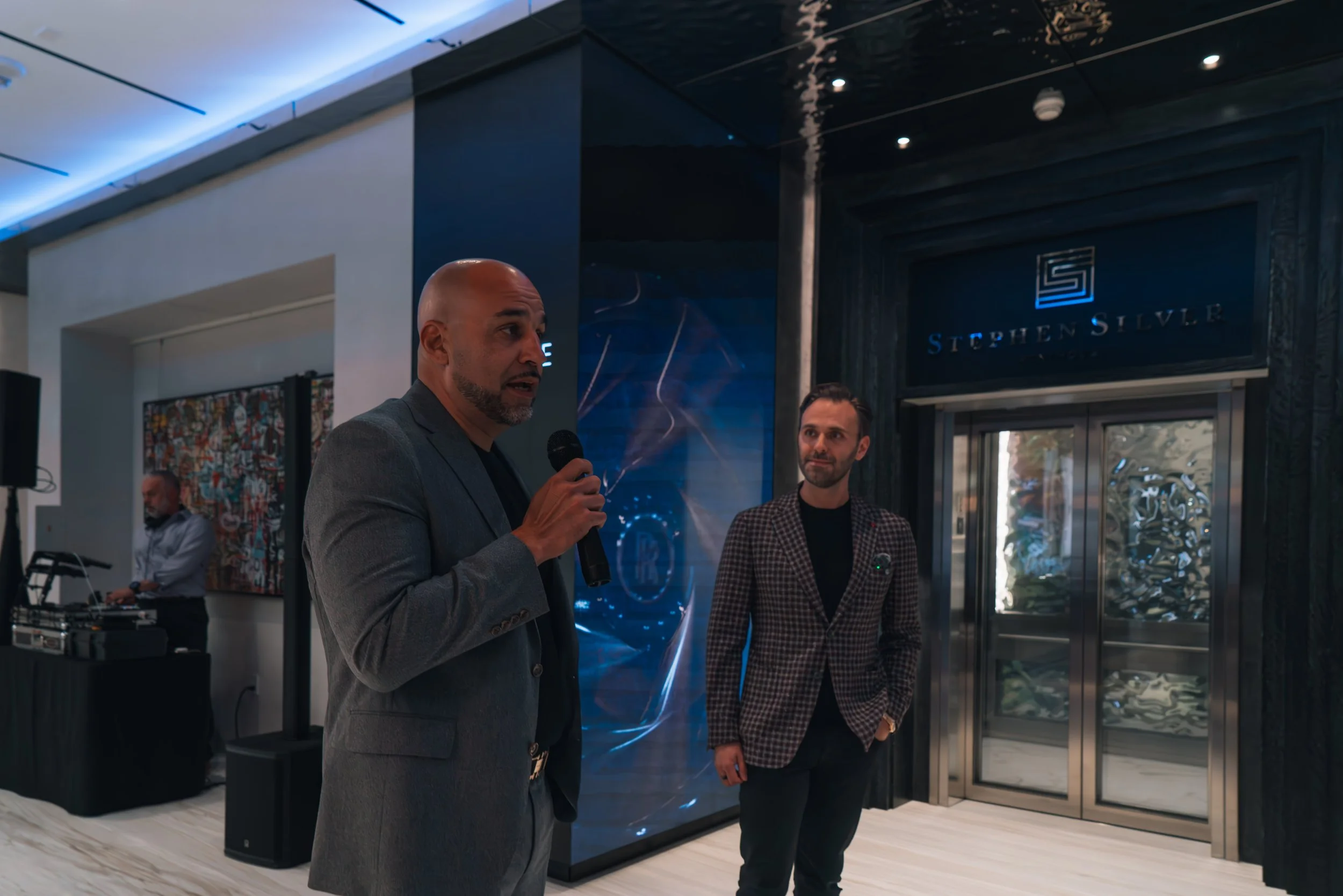 Two men in suits speaking at an event, one holding a microphone. One man has a shaved head and beard, the other has short dark hair. Behind them is a DJ setup and the entrance to Stephen Silver.