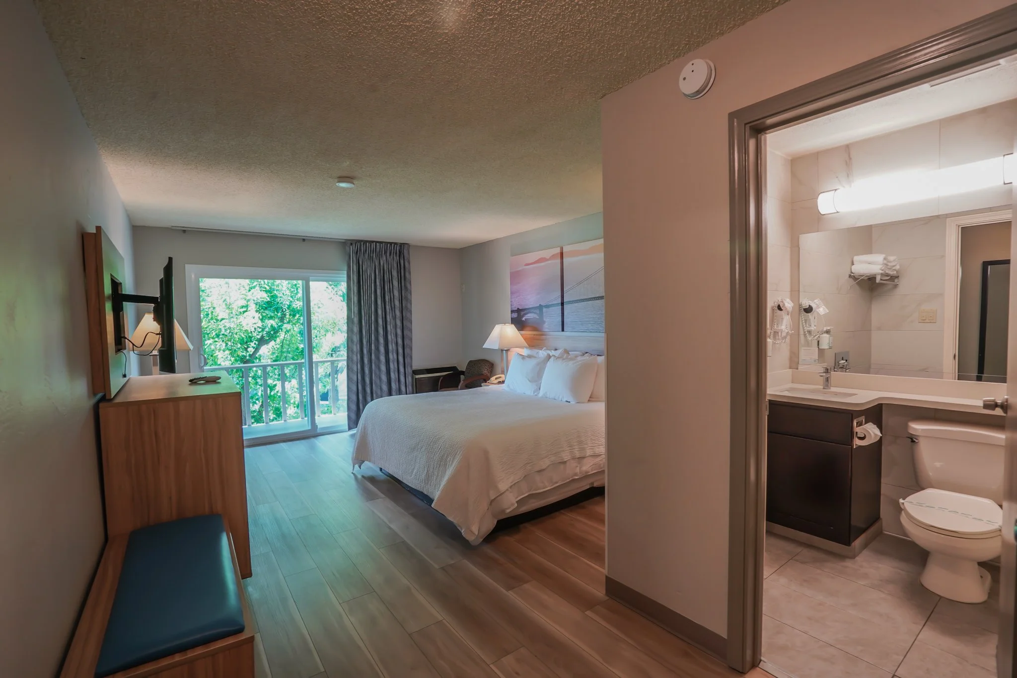 Hotel room with a large bed, balcony door with curtains, and a bathroom visible through an open door.