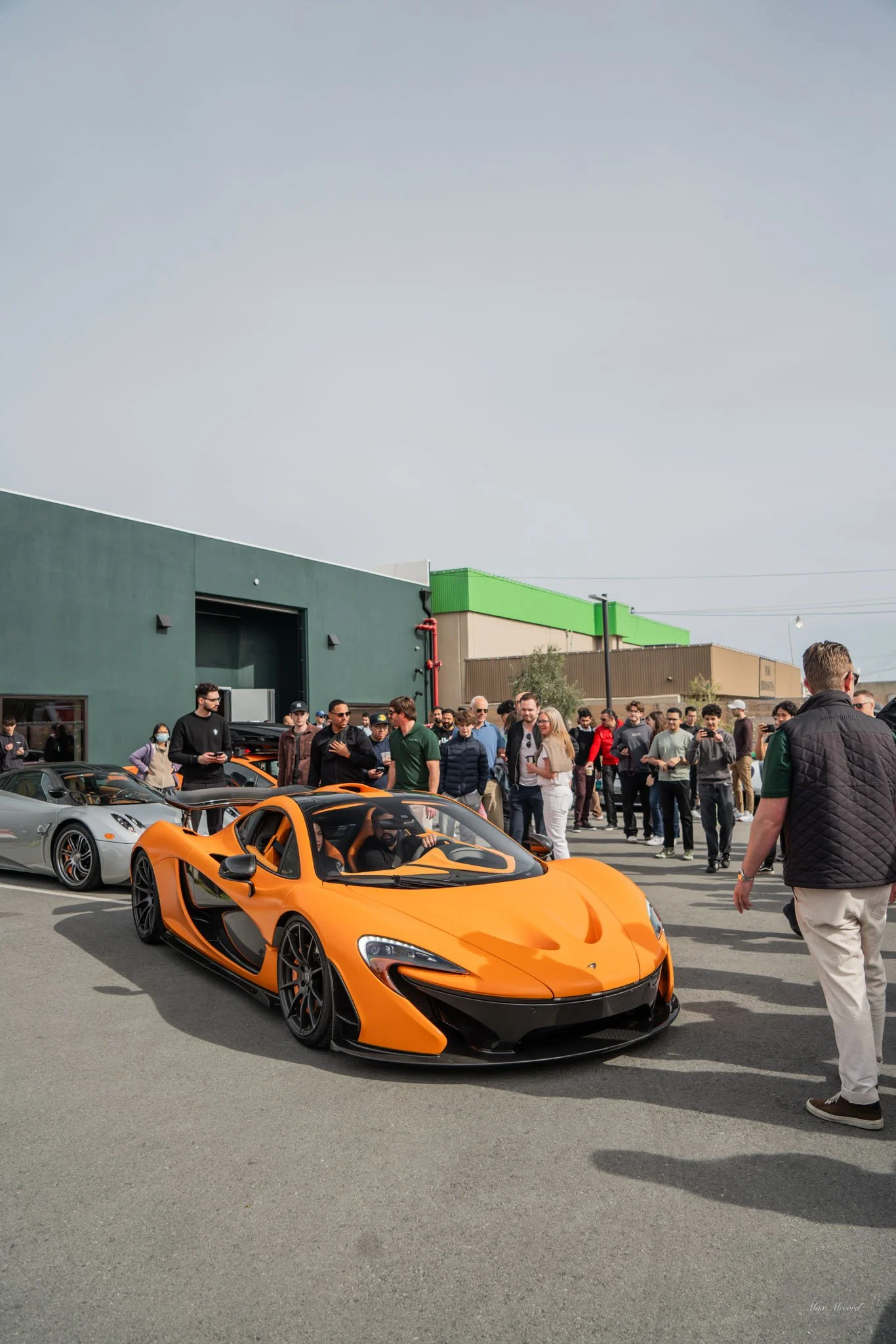An orange McLaren sports car parked among a crowd of people in an outdoor event, with some people taking photos and others observing the cars.