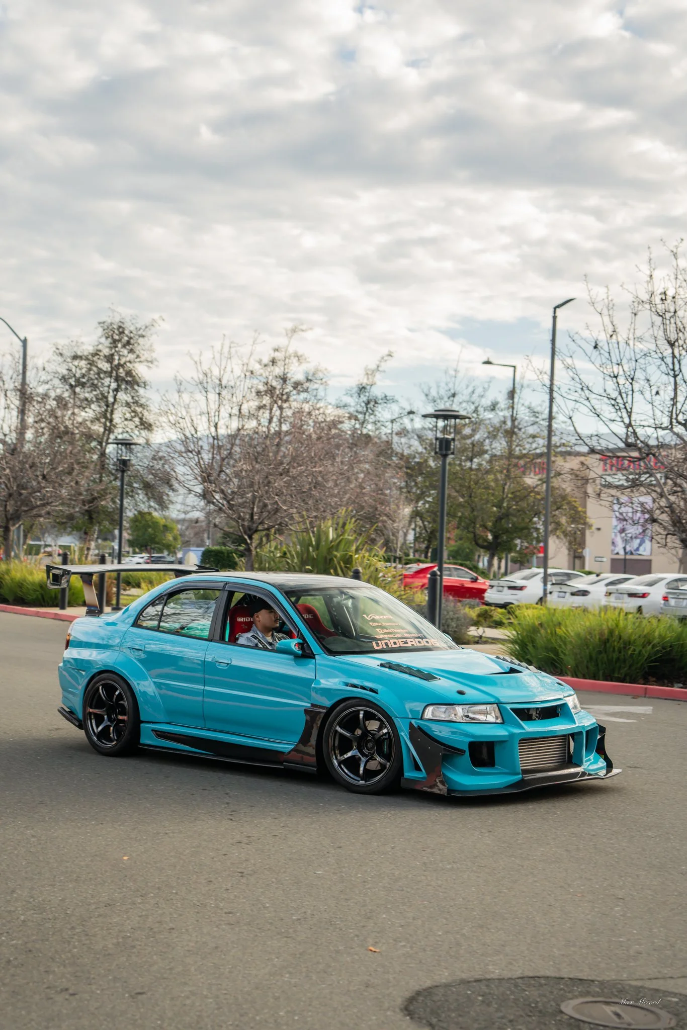A blue modified sports car with a large rear wing, black rims, and a front bumper with an intercooler, driving on a city street with trees and parked cars in the background.