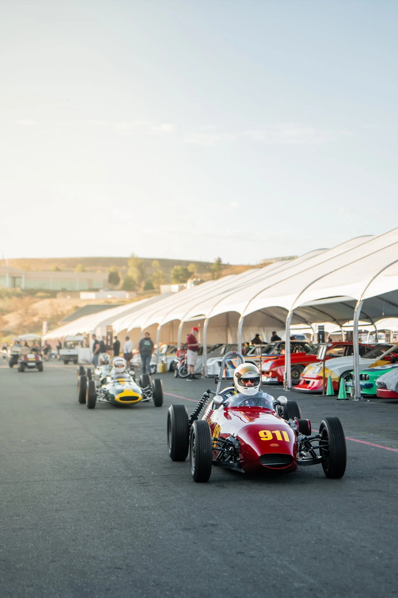 Vintage race cars lined up at a racing event with tents and spectators in the background.