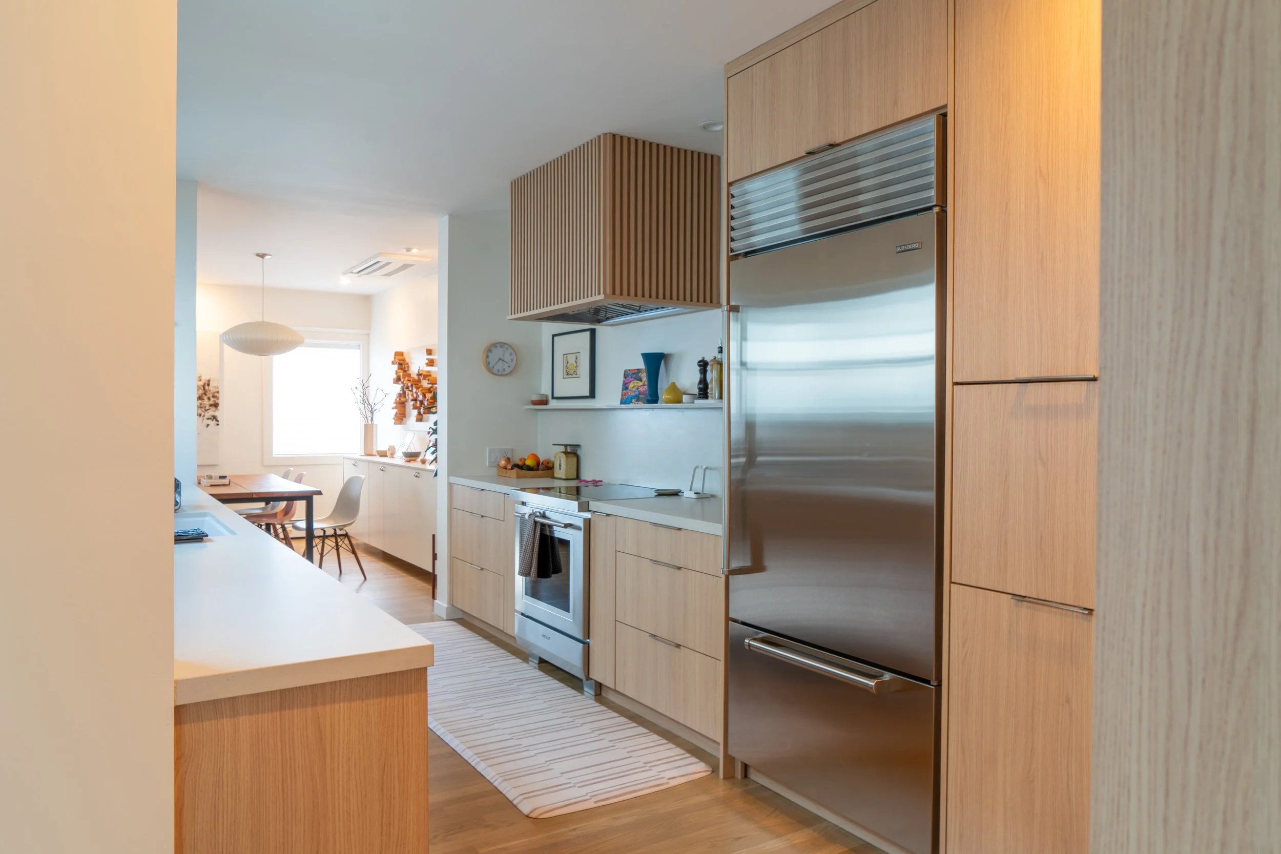 Modern kitchen with wooden cabinetry, stainless steel refrigerator, white countertops, and a dining area in the background with a white dining table and chairs.