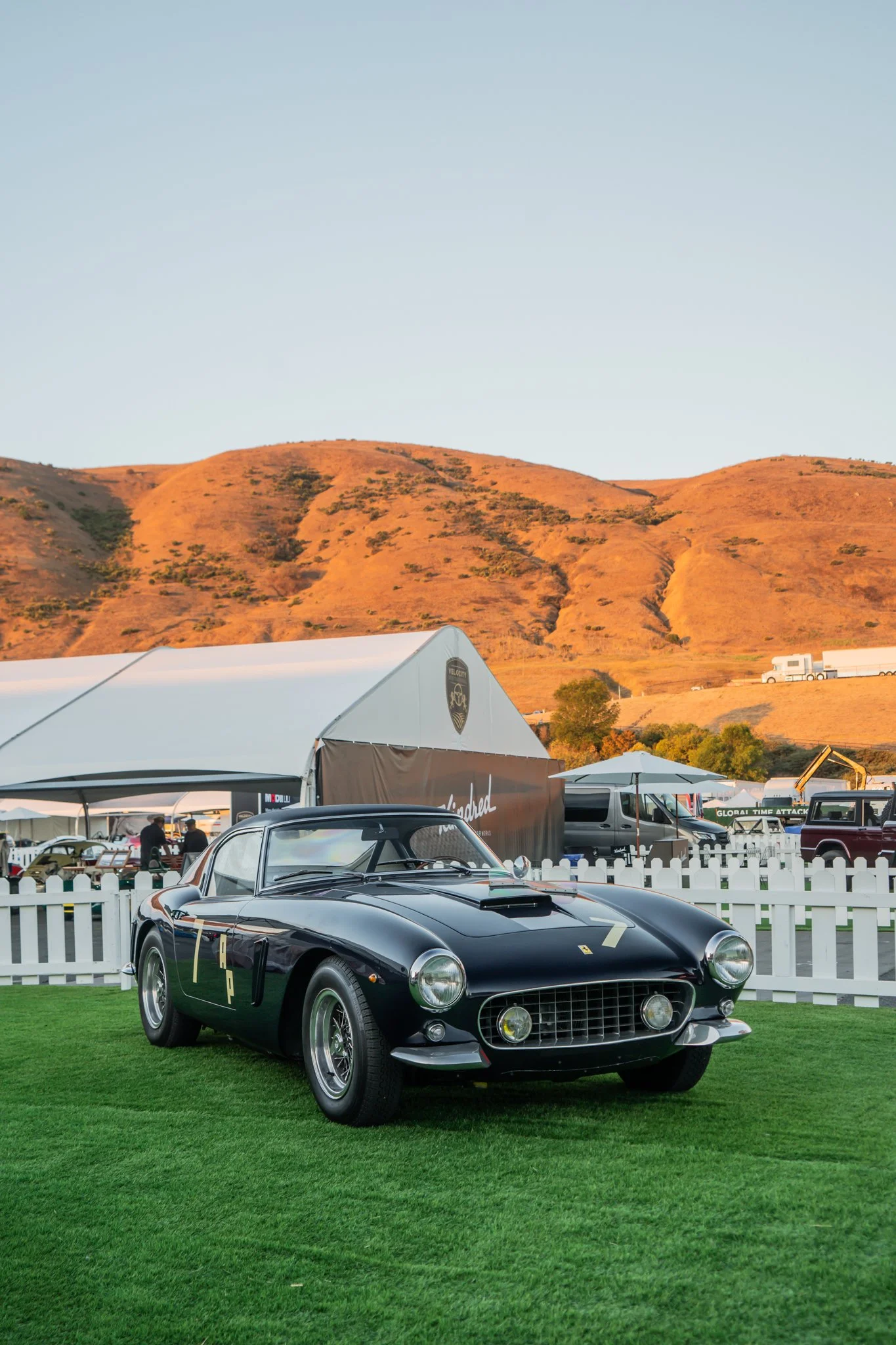 A black vintage Ferrari sports car with racing numbers parked on green grass at an outdoor car event, with tents and hills in the background.