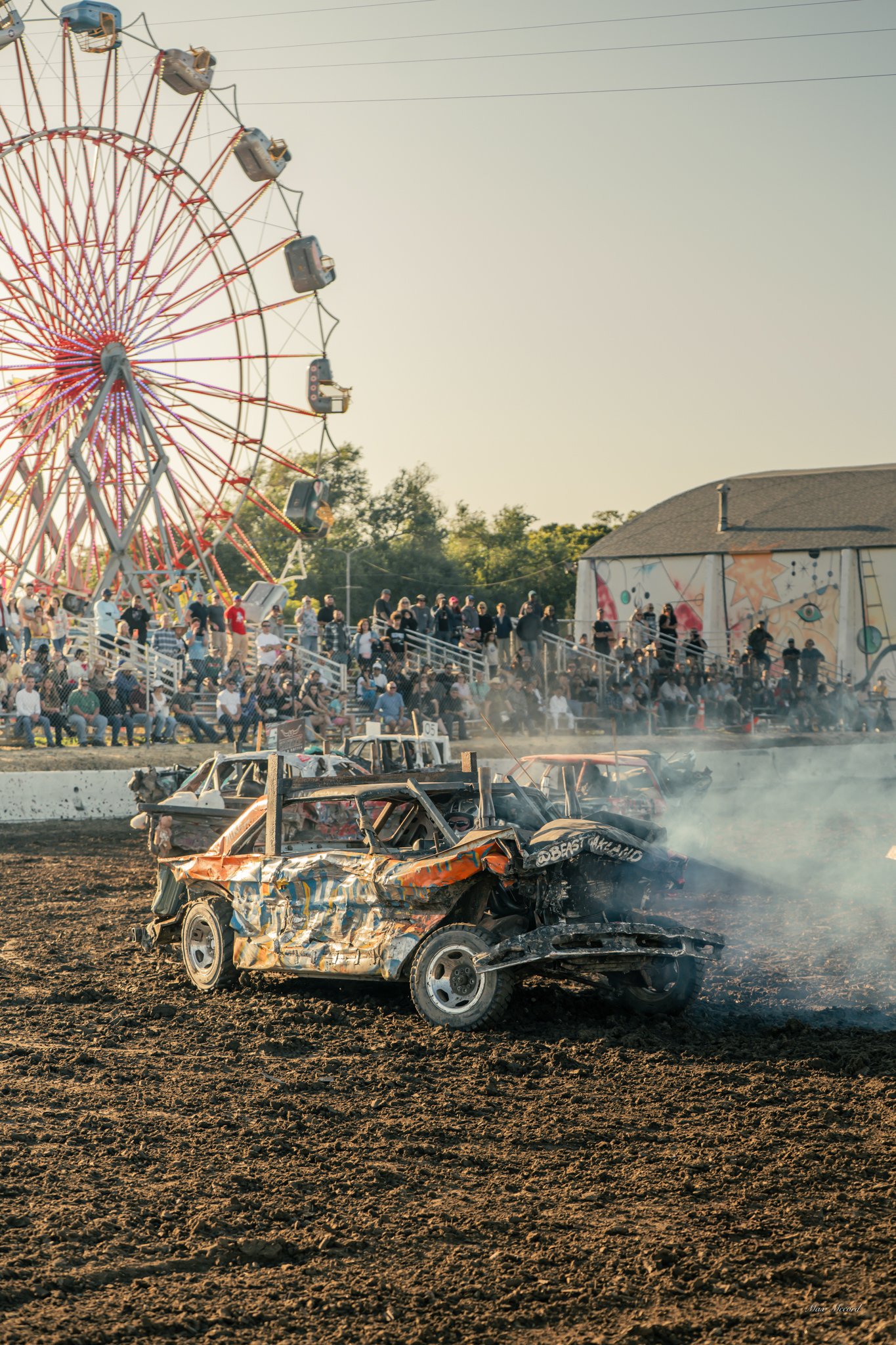 A wrecked car on a dirt track during a demolition derby at a fair with a ferris wheel and a crowd of spectators in the background.