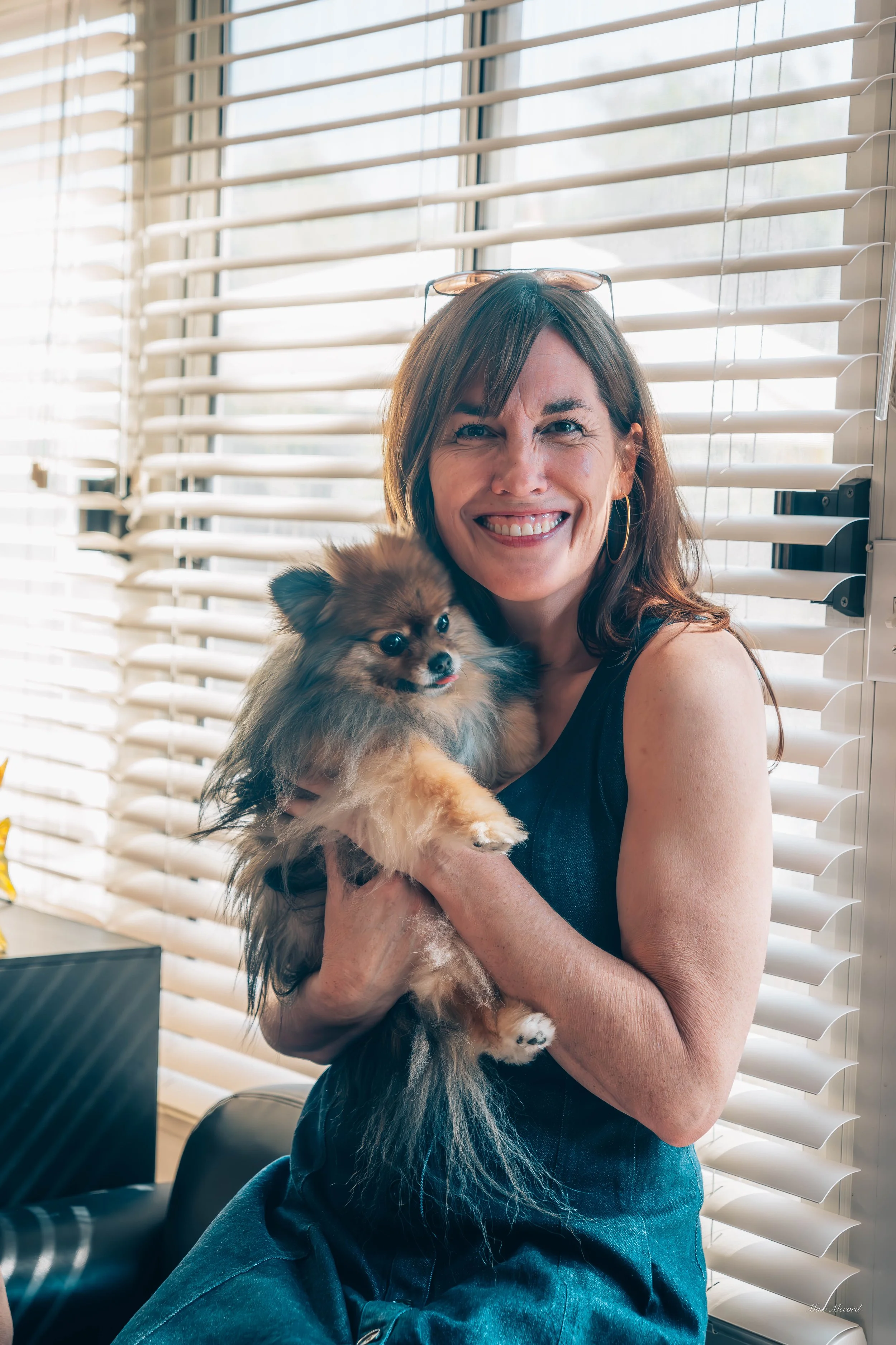 A woman with brown hair, wearing a sleeveless top and glasses on her head, holding a small fluffy dog near a window with white blinds.