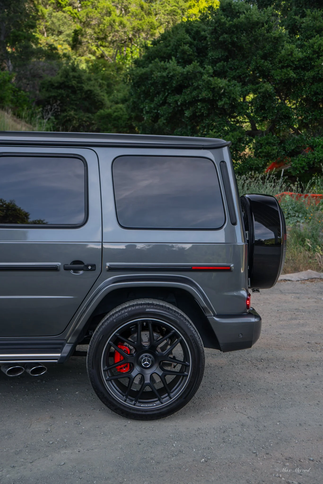 Side view of a gray Mercedes-Benz G-Class SUV parked on a dirt road, with green trees and vegetation in the background.