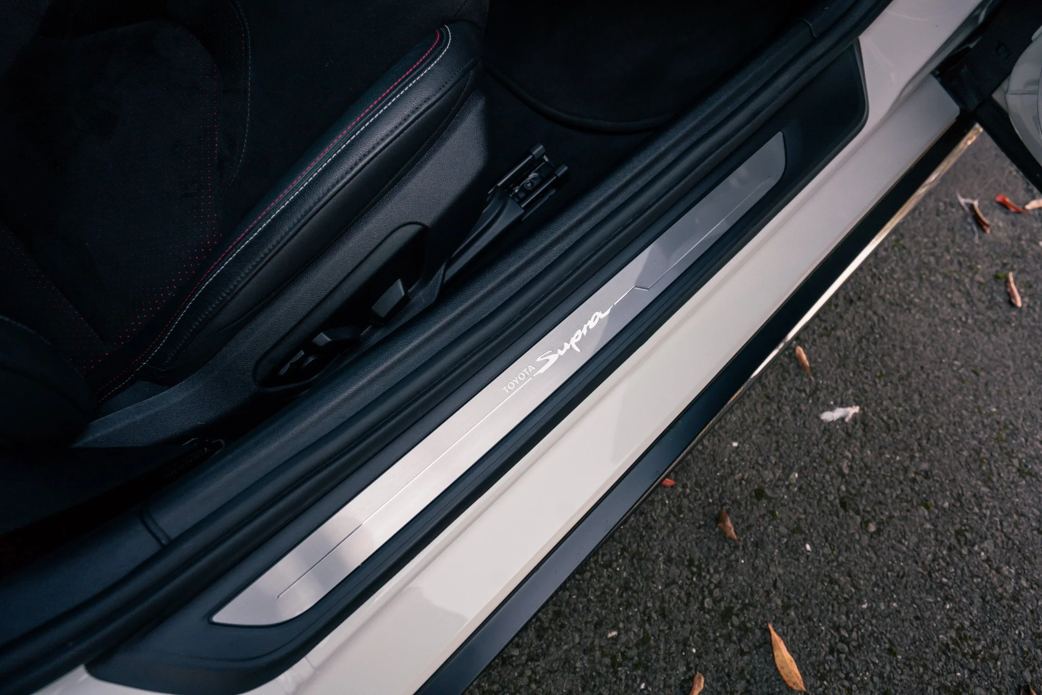 Close-up of the door sill of a white Toyota Supra, showing the black interior with red stitching and the pavement outside.
