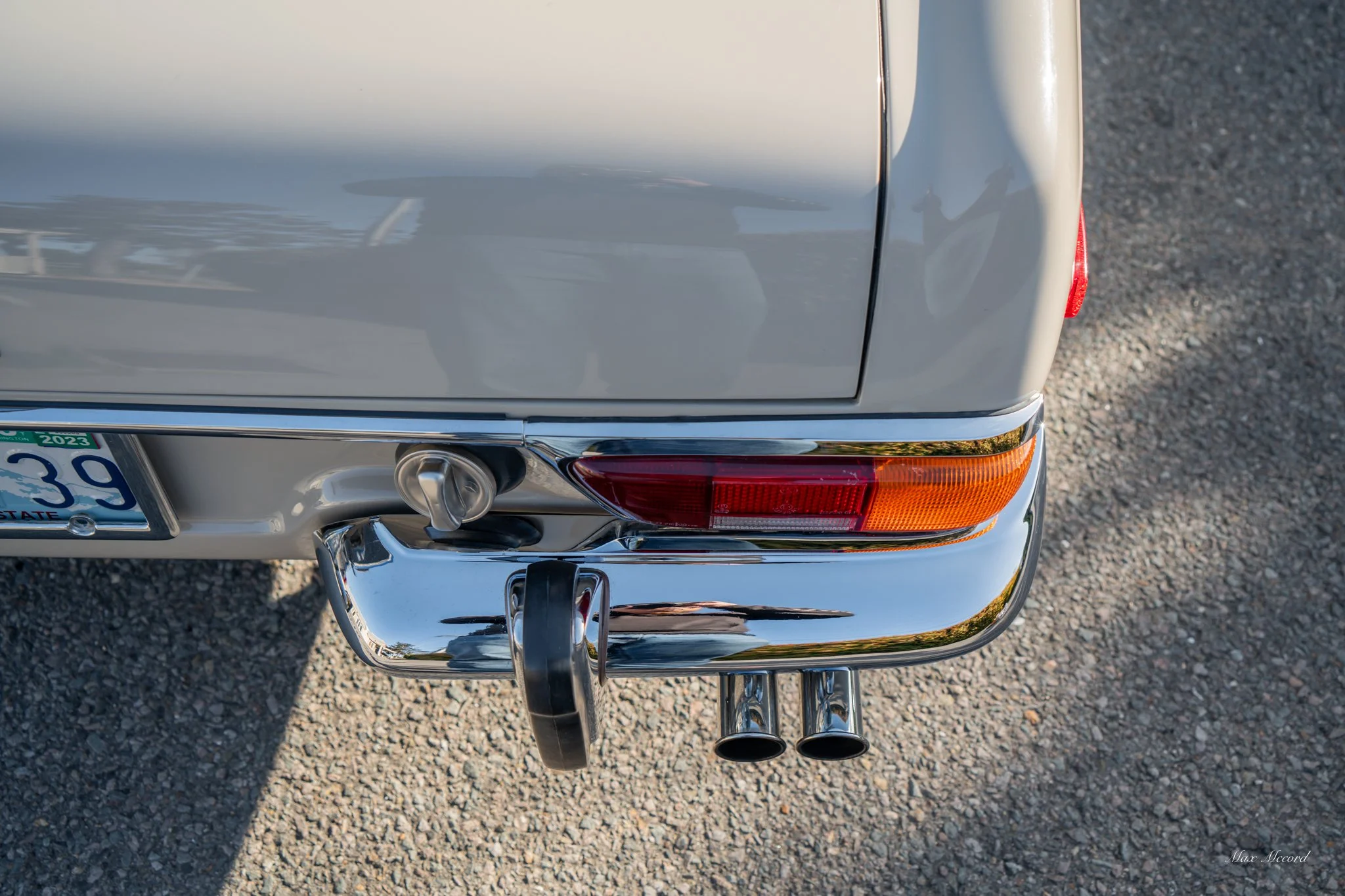 Close-up of the rear end of a vintage silver car, showing the bumper, tail light, dual exhaust pipes, and a partially visible license plate.