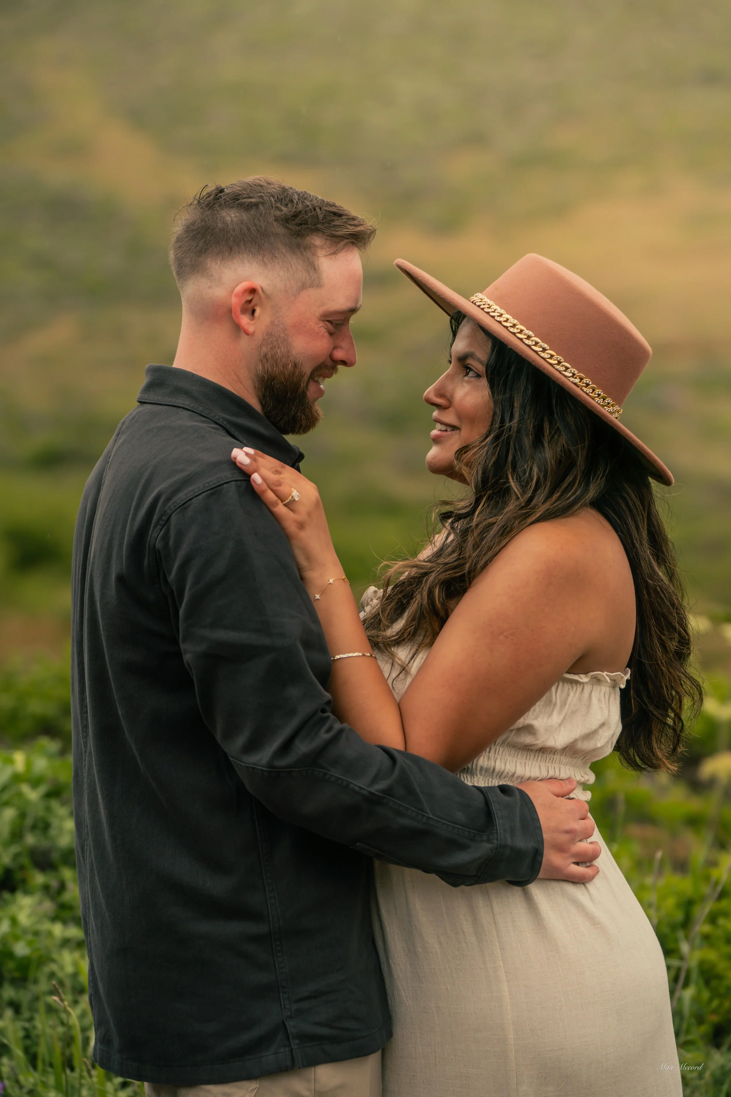 A couple standing close together outdoors, smiling at each other, with a blurred green natural background. The woman is wearing a tan wide-brimmed hat and a sleeveless dress, and the man is wearing a dark shirt.