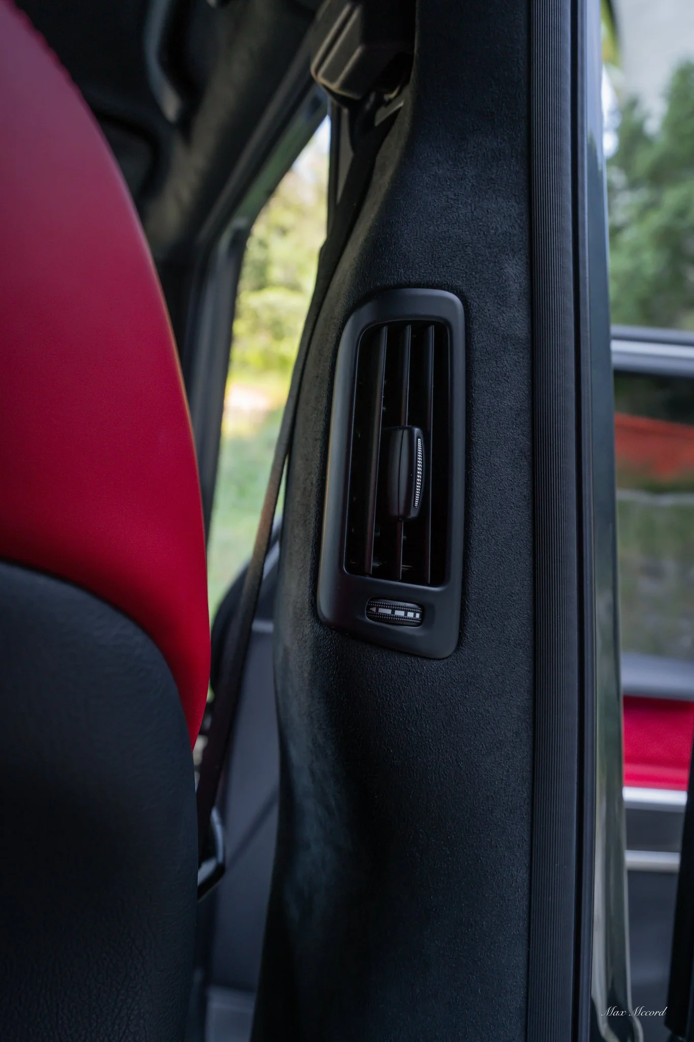 Interior of a vehicle showing a black air conditioning vent on the side of the front passenger seat with a red seat in the background.