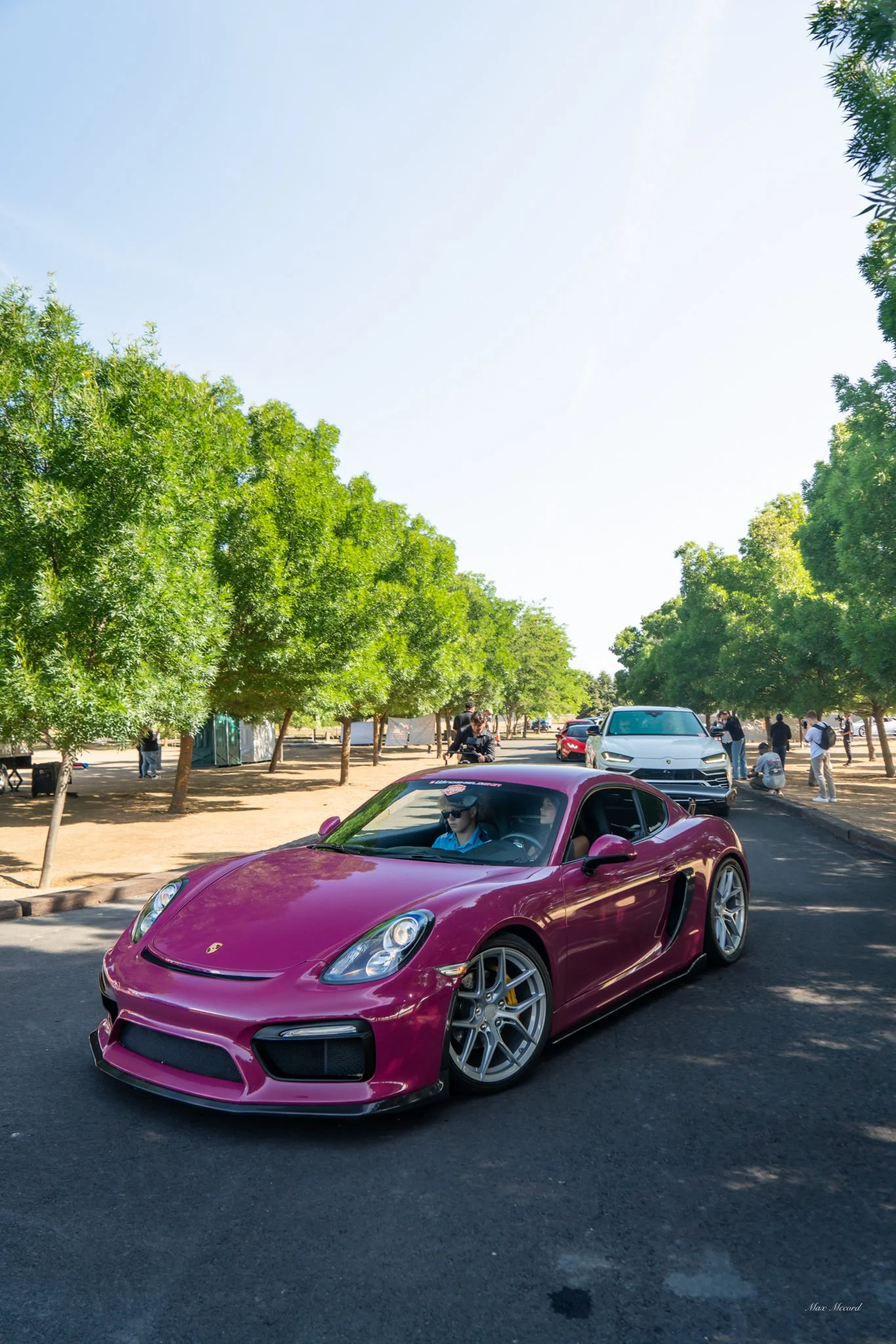 A maroon Porsche sports car driving on the road, surrounded by green trees and other cars, with people walking on the side.