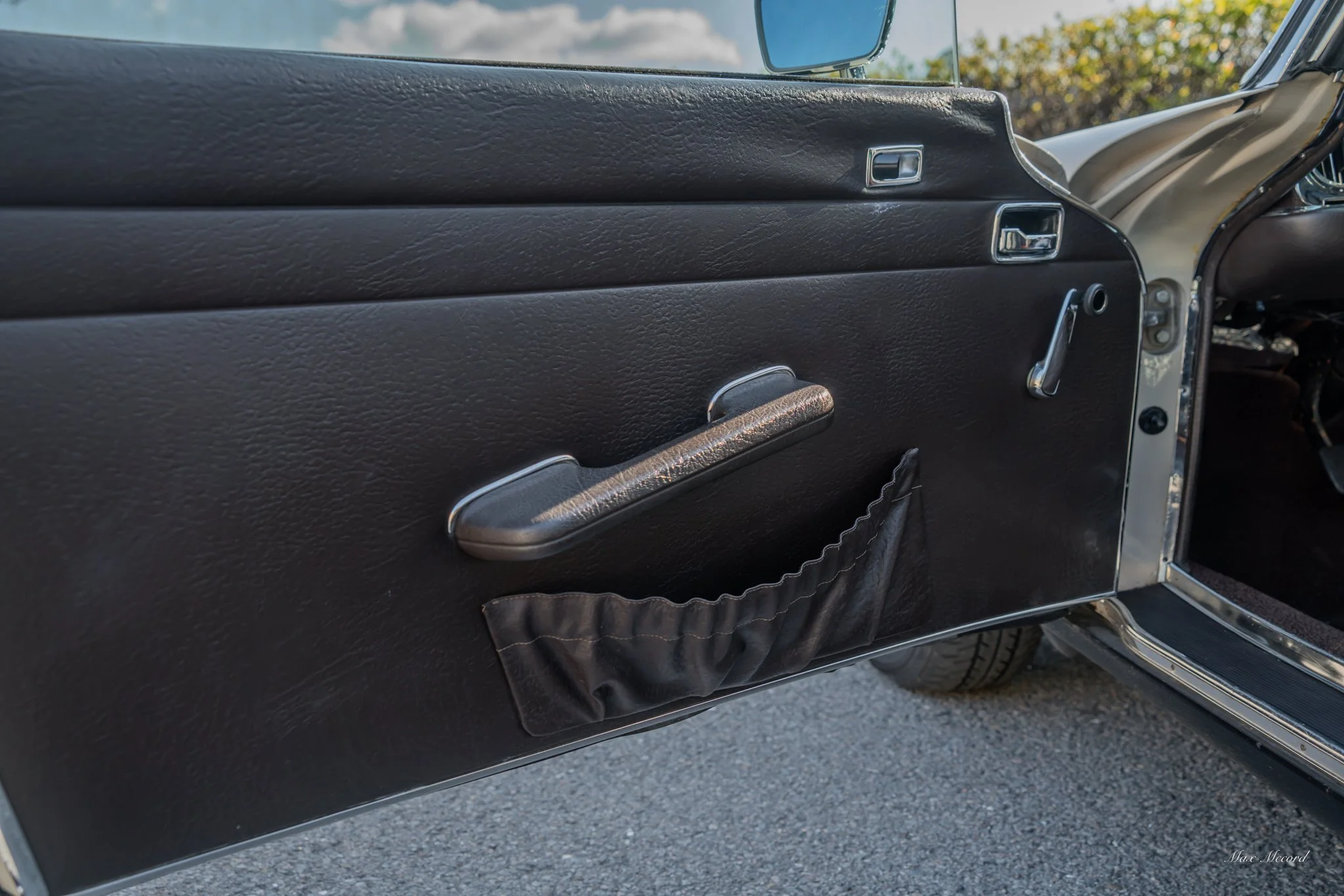 The inside of a vintage car door with a black textured surface, a manual window crank handle, a black leather storage pouch, and a side mirror visible through the window.