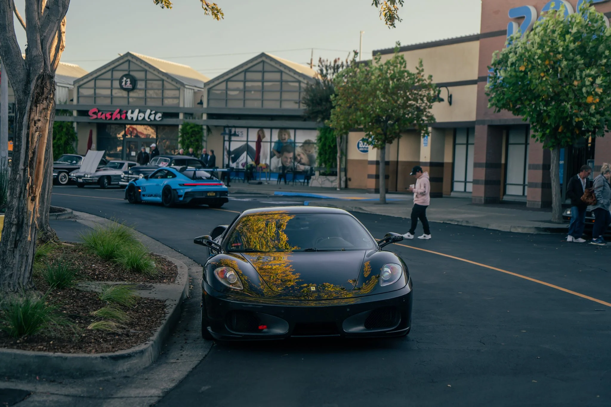 A black sports car parked on the street with trees and a shopping center in the background. Multiple other cars are parked nearby, and a person is walking on the sidewalk looking at their phone.