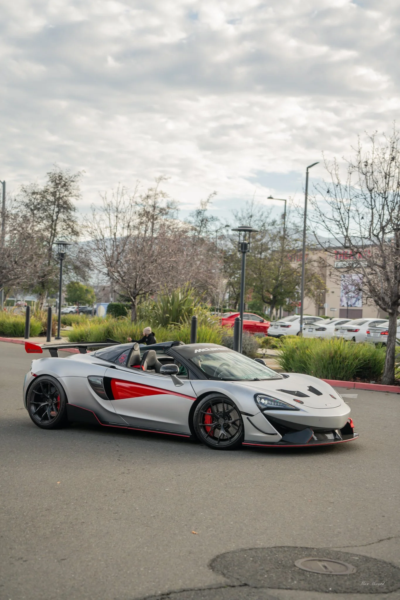 A silver and black McLaren race car with red accents parked in a lot with leafless trees and a cloudy sky in the background.