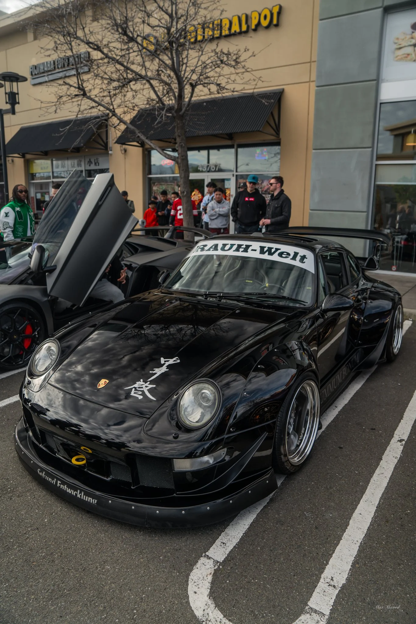 A black Porsche race car with Japanese characters on the hood, parked at an outdoor event, with a group of people in the background near storefronts.