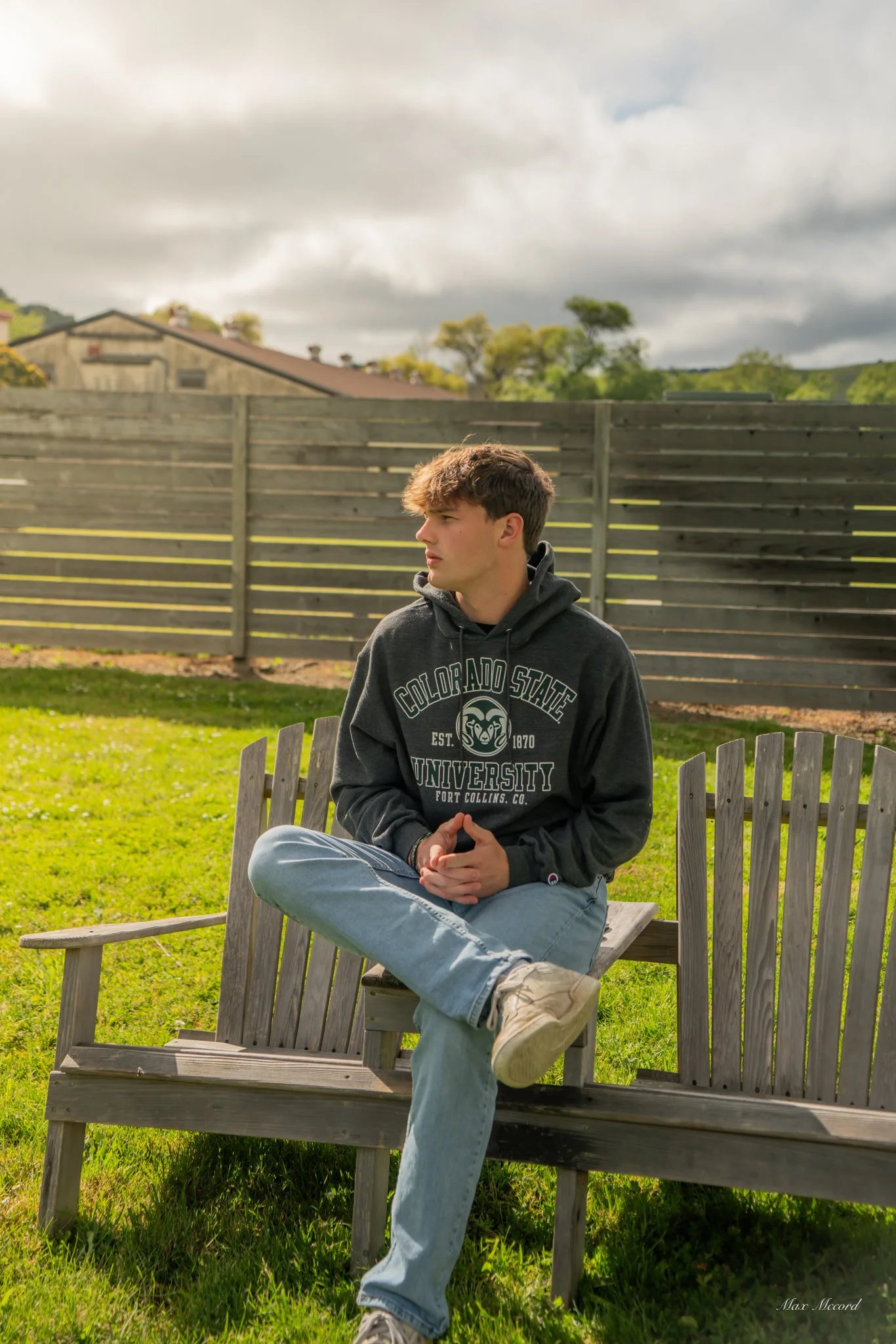 A young man with light skin and brown hair is sitting cross-legged on a wooden bench outdoors. He is wearing a dark gray hoodie with white text and graphics, light blue jeans, and beige sneakers. He is looking to his left with a neutral expression, a