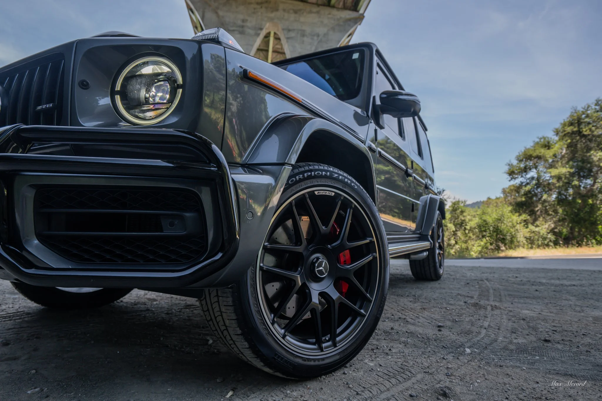 Close-up of a black Mercedes-Benz G-Class SUV from a low angle, showing the front left side with black wheels and red brake calipers, parked outdoors near a bridge and trees.