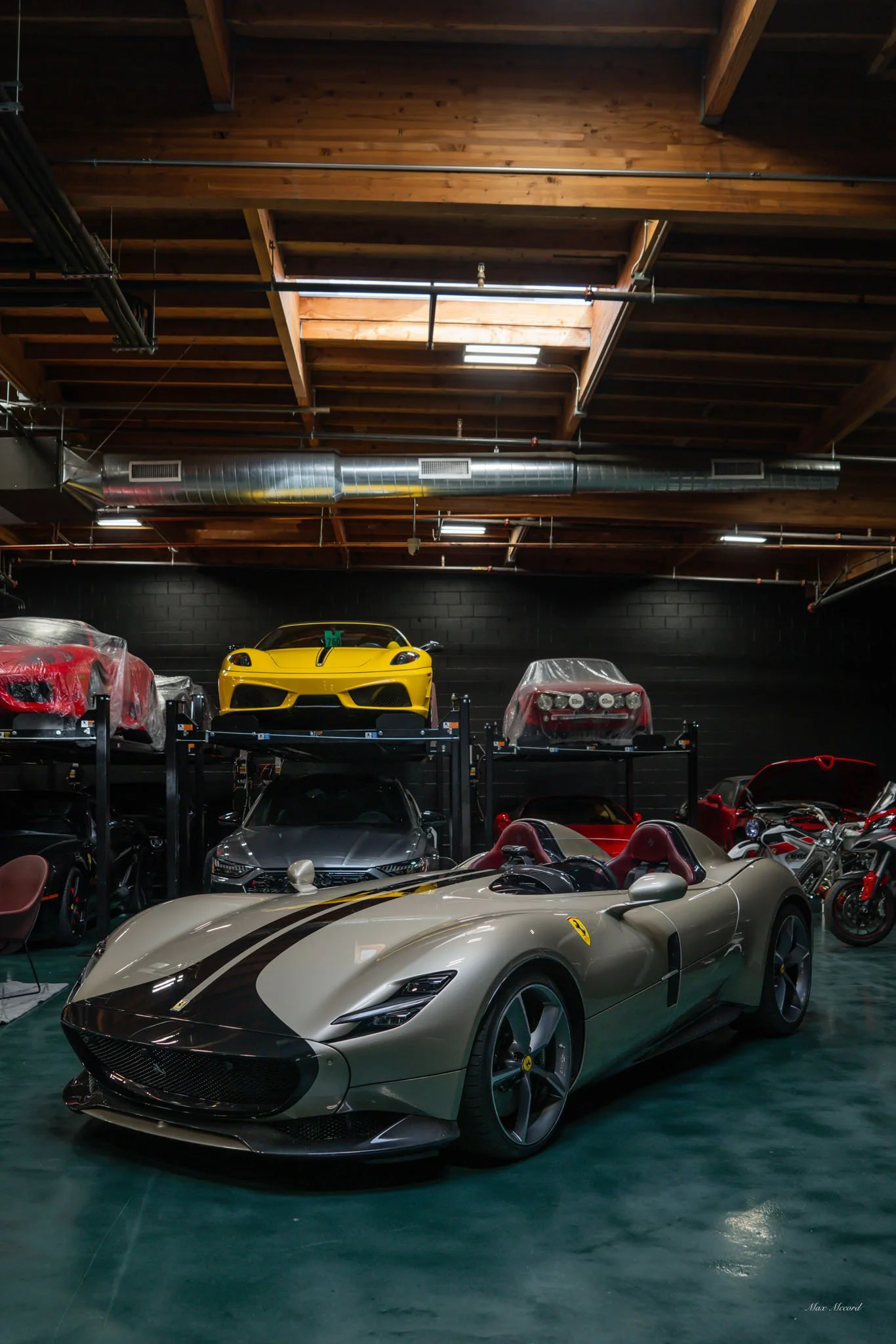 A luxury sports car garage with a beige Ferrari in the foreground and various high-end cars, including a yellow Ferrari, stored on elevated racks in the background.