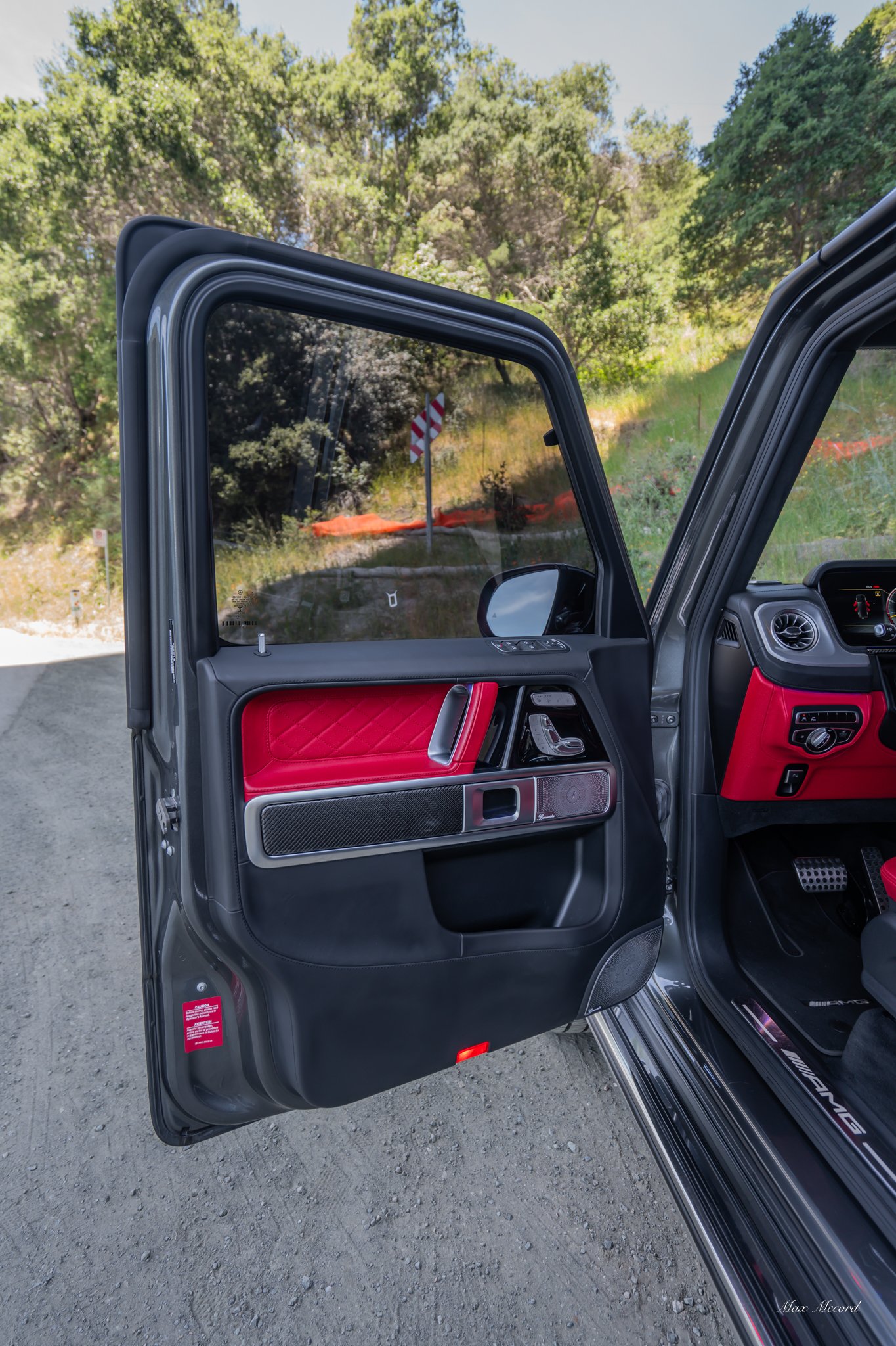 Open car door showing black and red interior with carbon fiber trim and a seat with AMG branding, on a dirt road with trees and a guardrail outside.