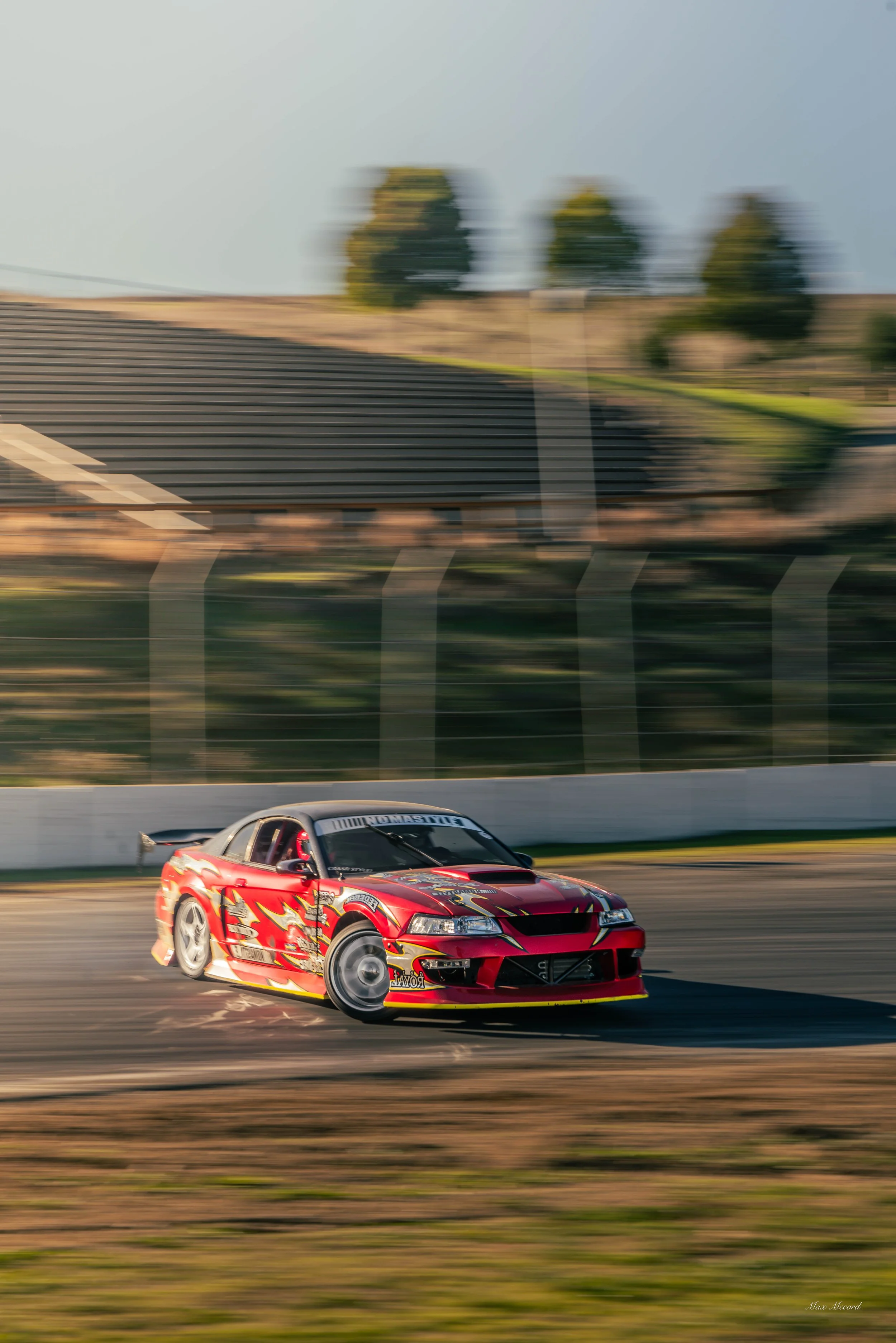 A red race car with sponsor logos speeds around a racetrack during daytime, with motion blur in the background.