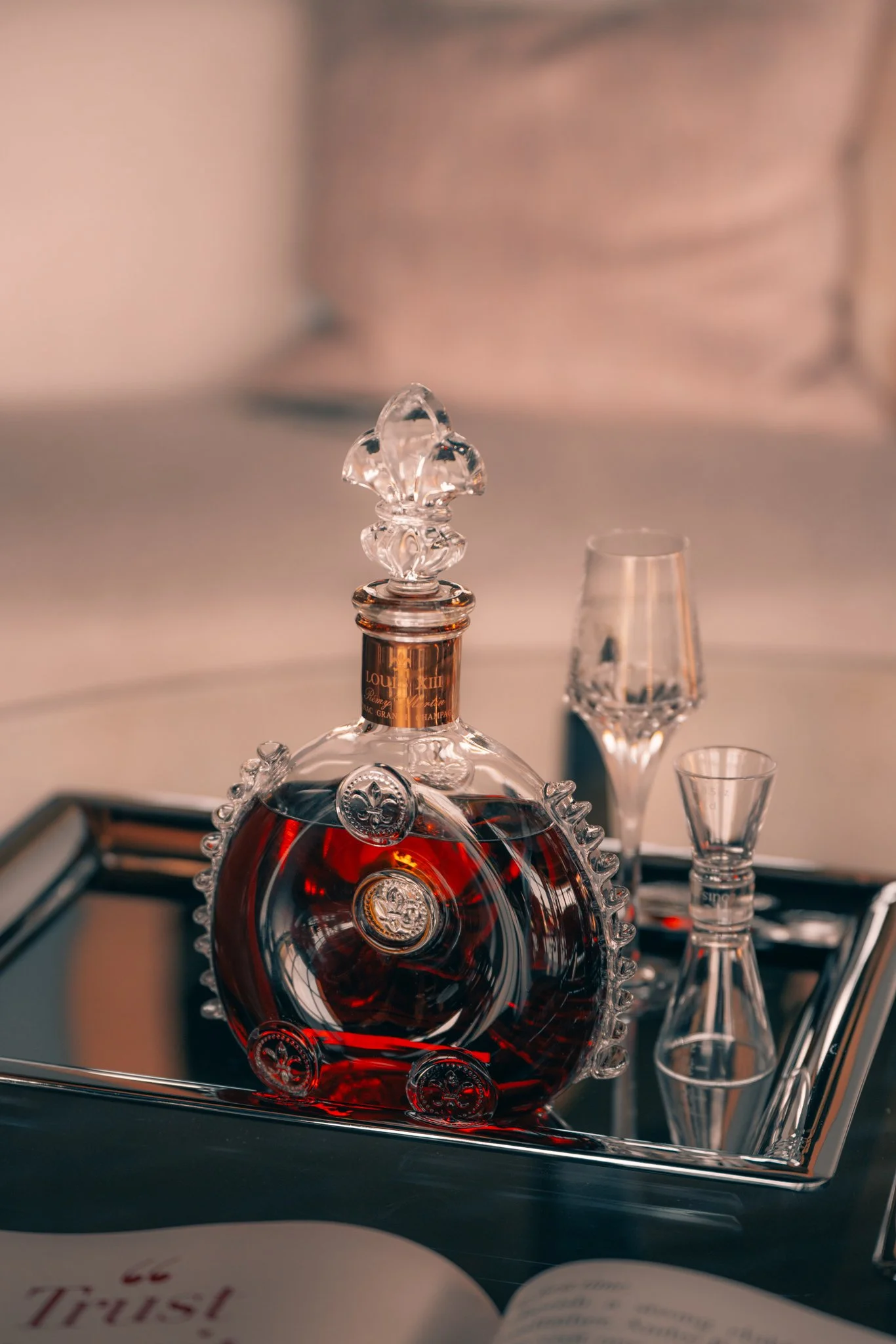 A decorative glass bottle filled with dark liquid, placed on a metal tray along with two small glasses, with a mirror reflecting the scene.