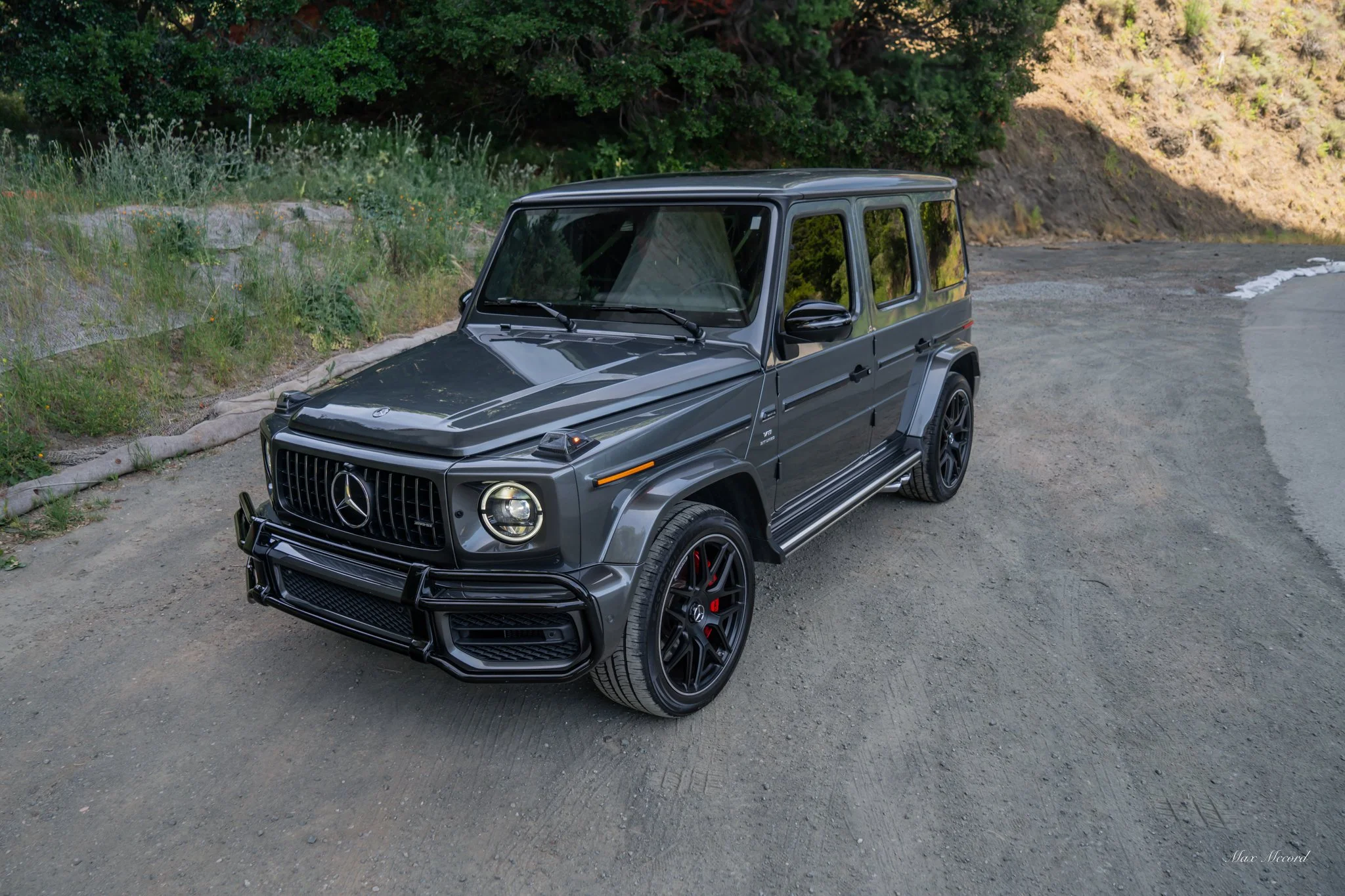 A black Mercedes-Benz G-Class SUV parked on a dirt roadside with greenery and hills in the background.