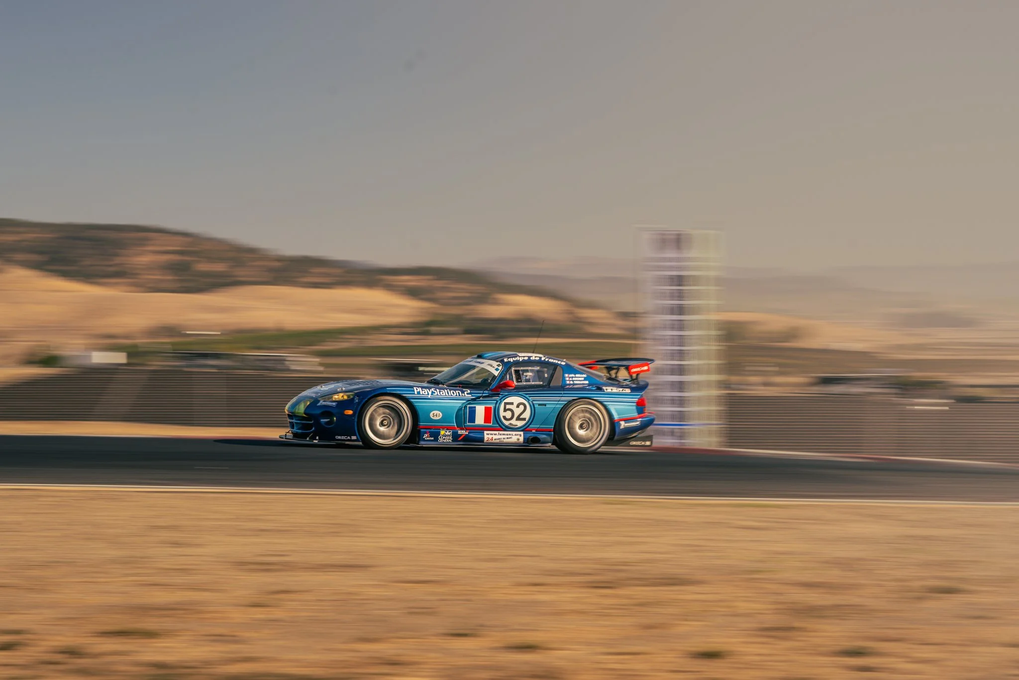 A blue race car with the number 52 is speeding on a race track, with a blurred background of hills and race barriers.