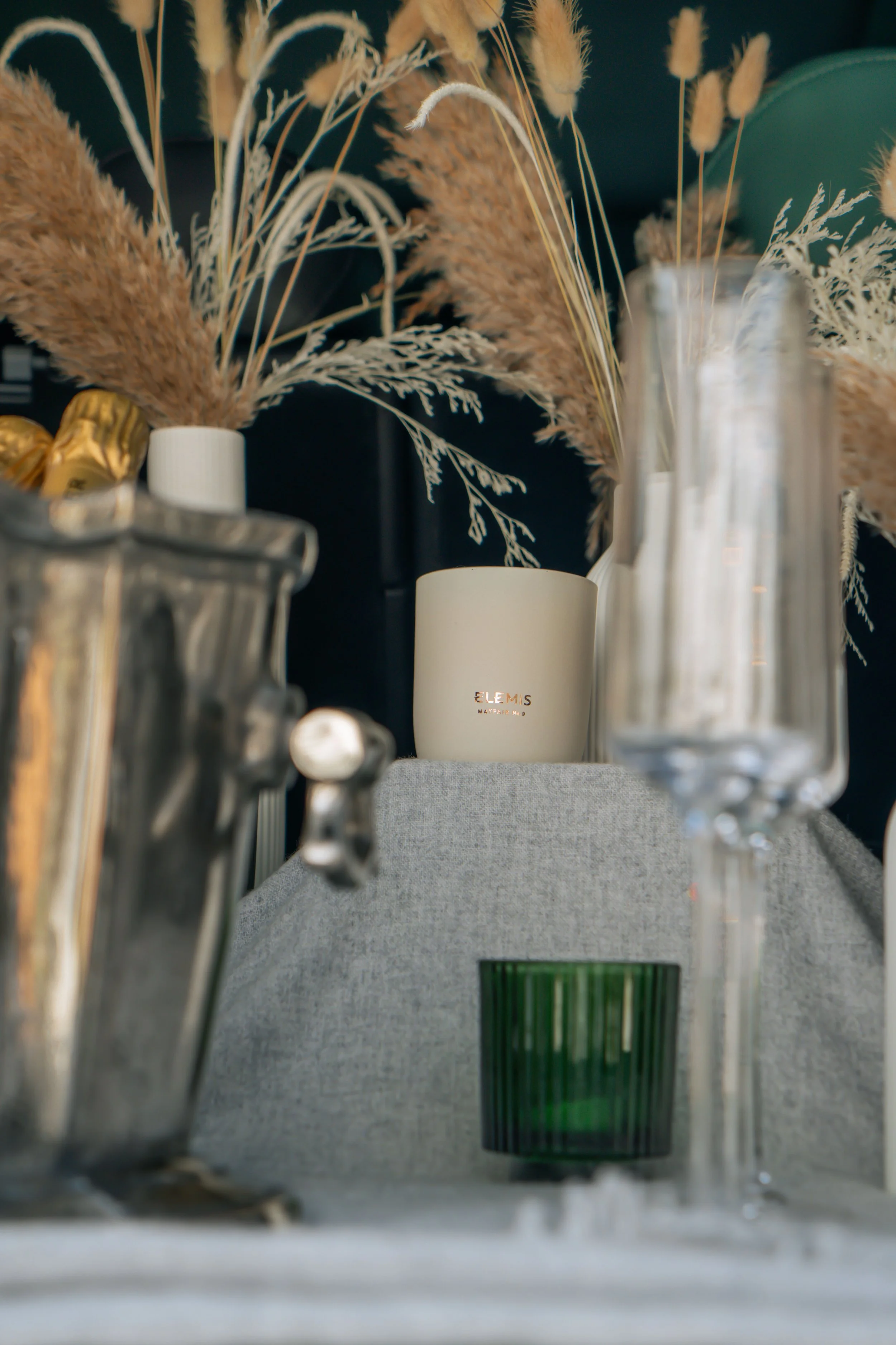 Decorative table setting featuring dried pampas grass and other plants in vases, glassware, and a candle.