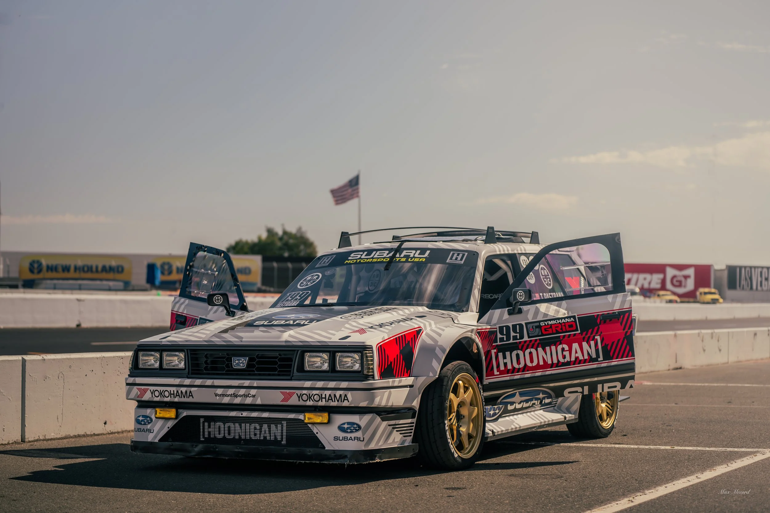 A vintage Subaru race car with a white, black, and red livery, gold wheels, and racing decals, parked on a racetrack with an American flag and sponsor banners in the background.