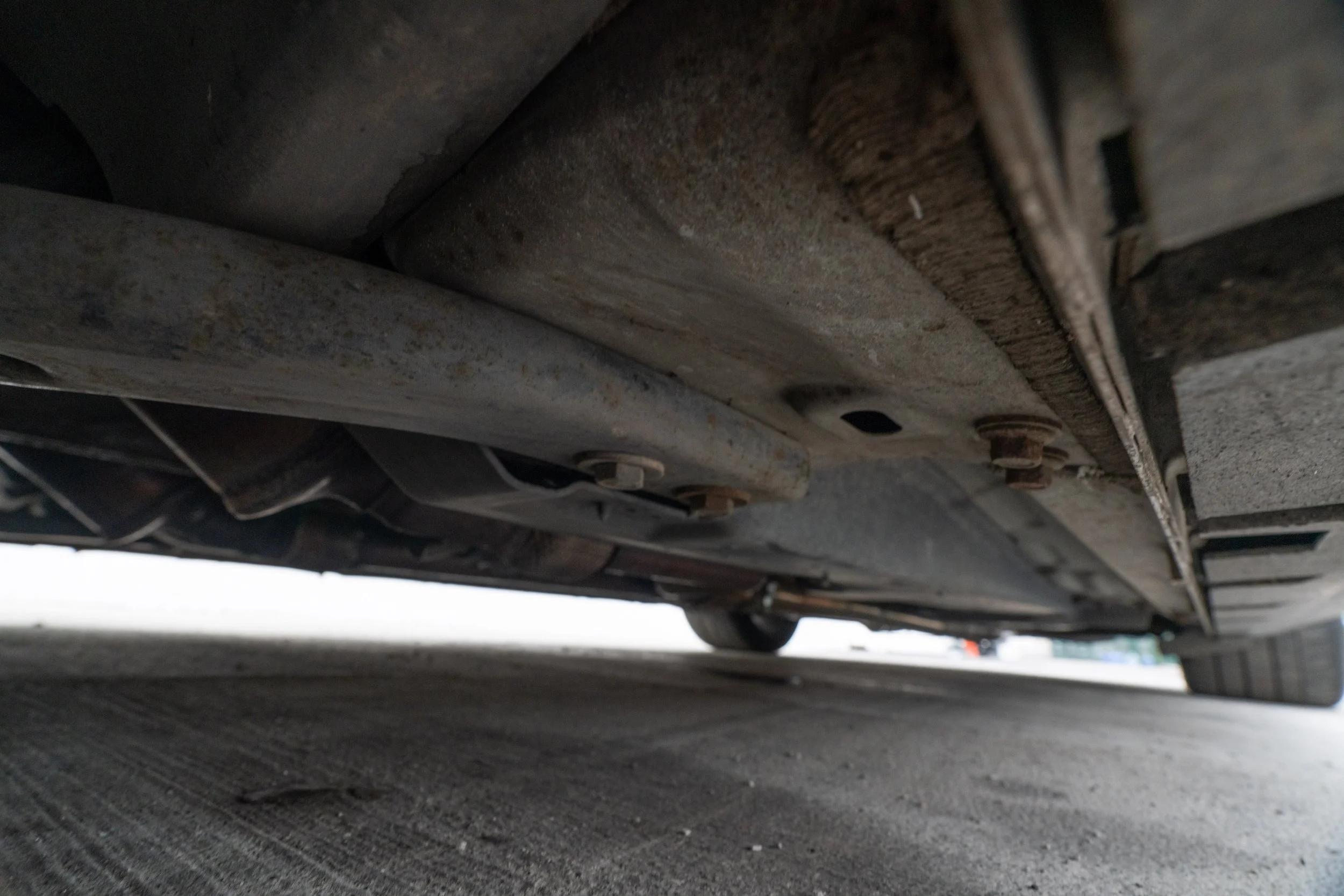 Underneath view of a car showing the underside with metal components, bolts, and rust, on a concrete surface.
